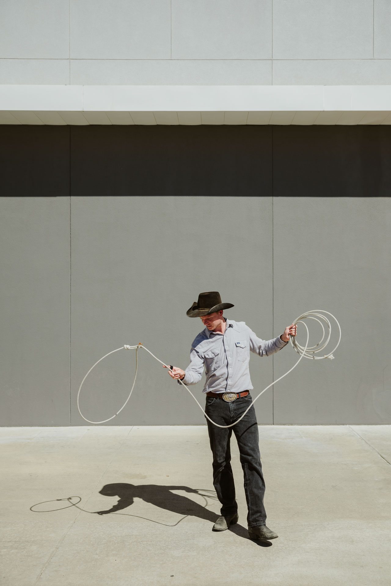 A man in a gray shirt with a black hat and rope performs a trick in front of a gray background