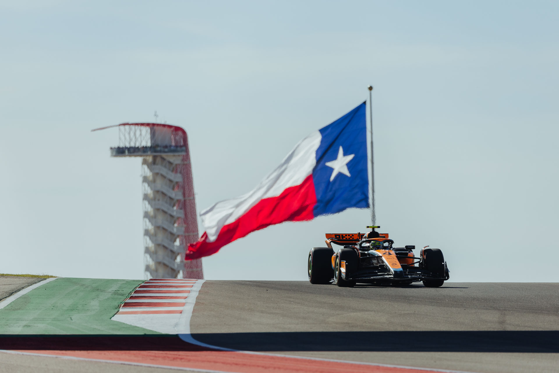 An F1 car passes in front of a large Texas flag above a strip of pavement