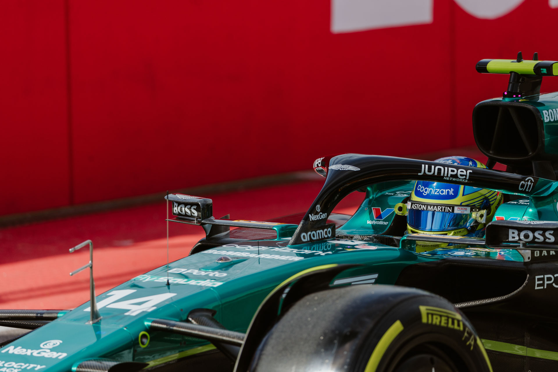 A detail shot of a green F1 car on a red background with a driver's helmet barely visible in the bottom right
