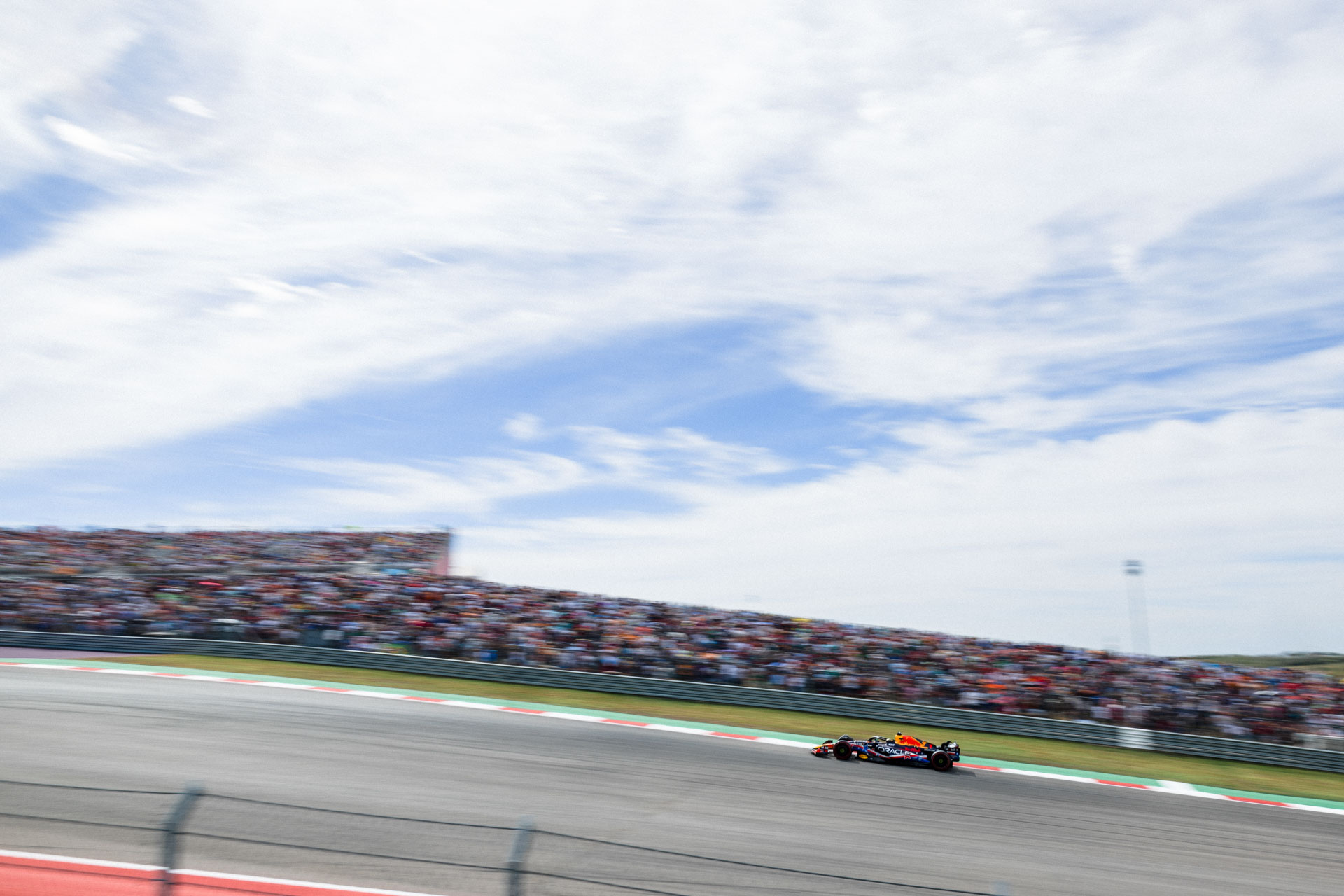 A racecar zooms past a blurred crowd under blue sky with white clouds