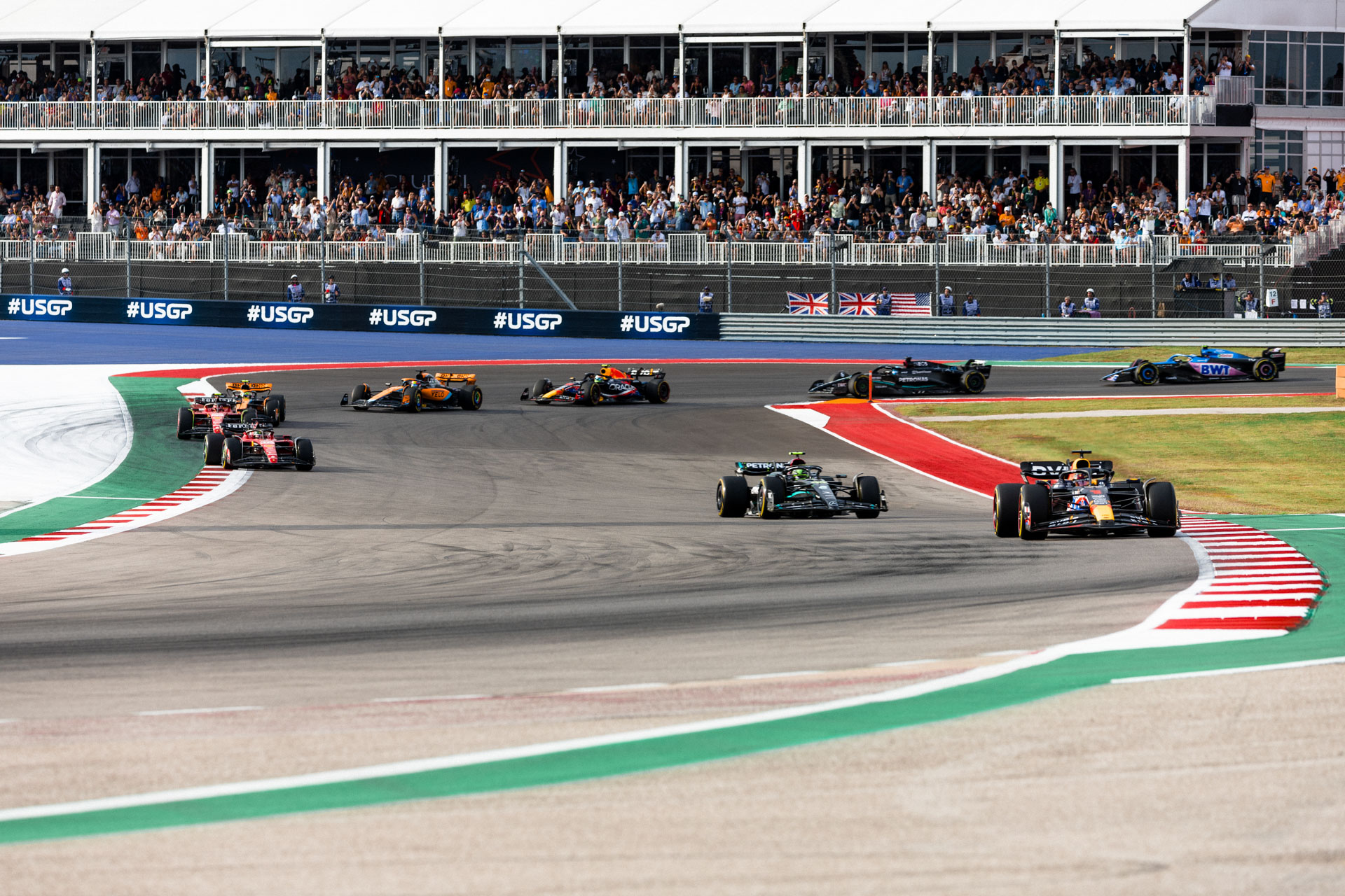 Eight F1 cars circle a pavement track painted in red, white, and green while onlookers view from shaded grandstands behind