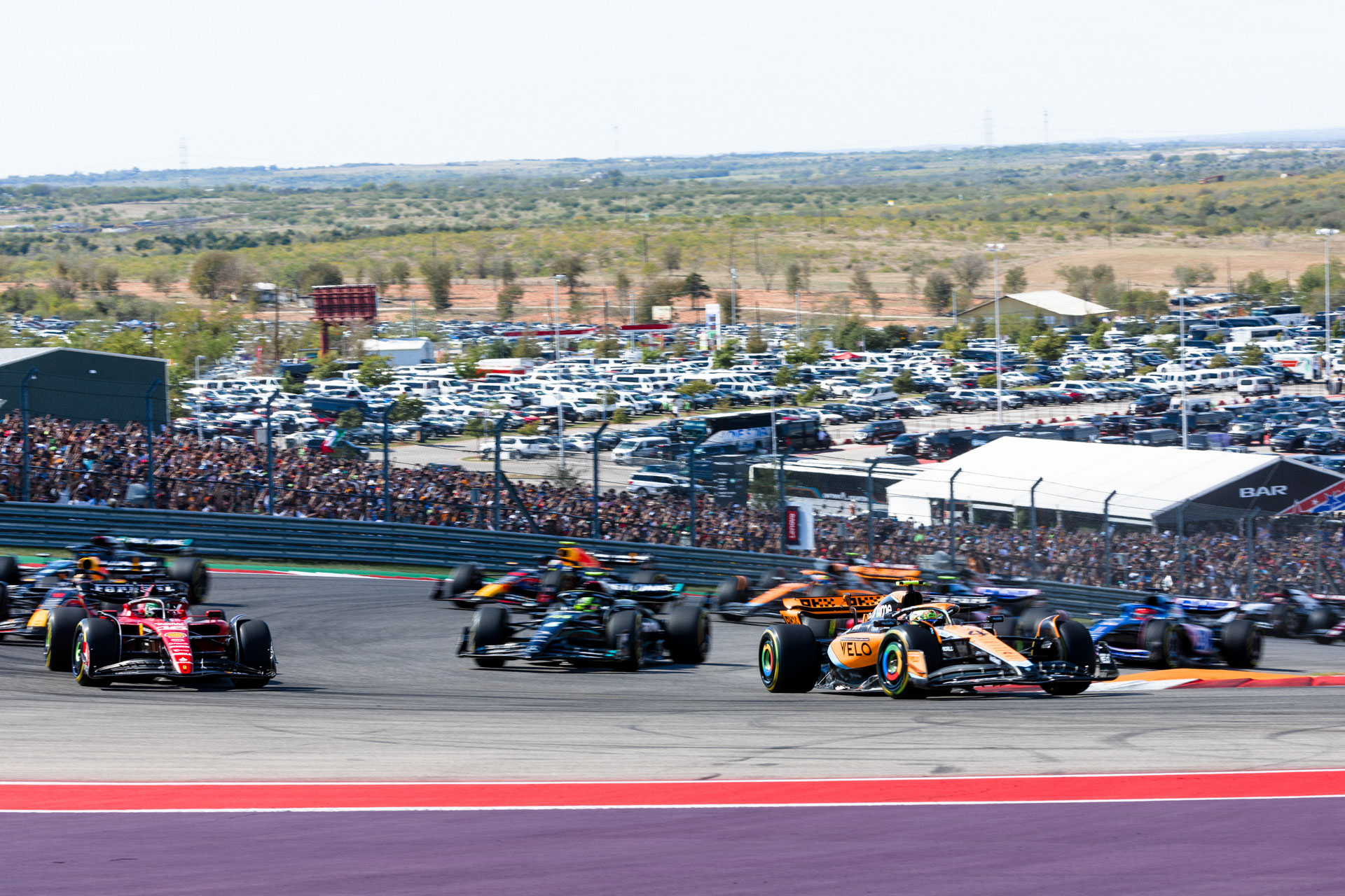 A group of F1 cars circle a pavement track while crowds watch in the distance