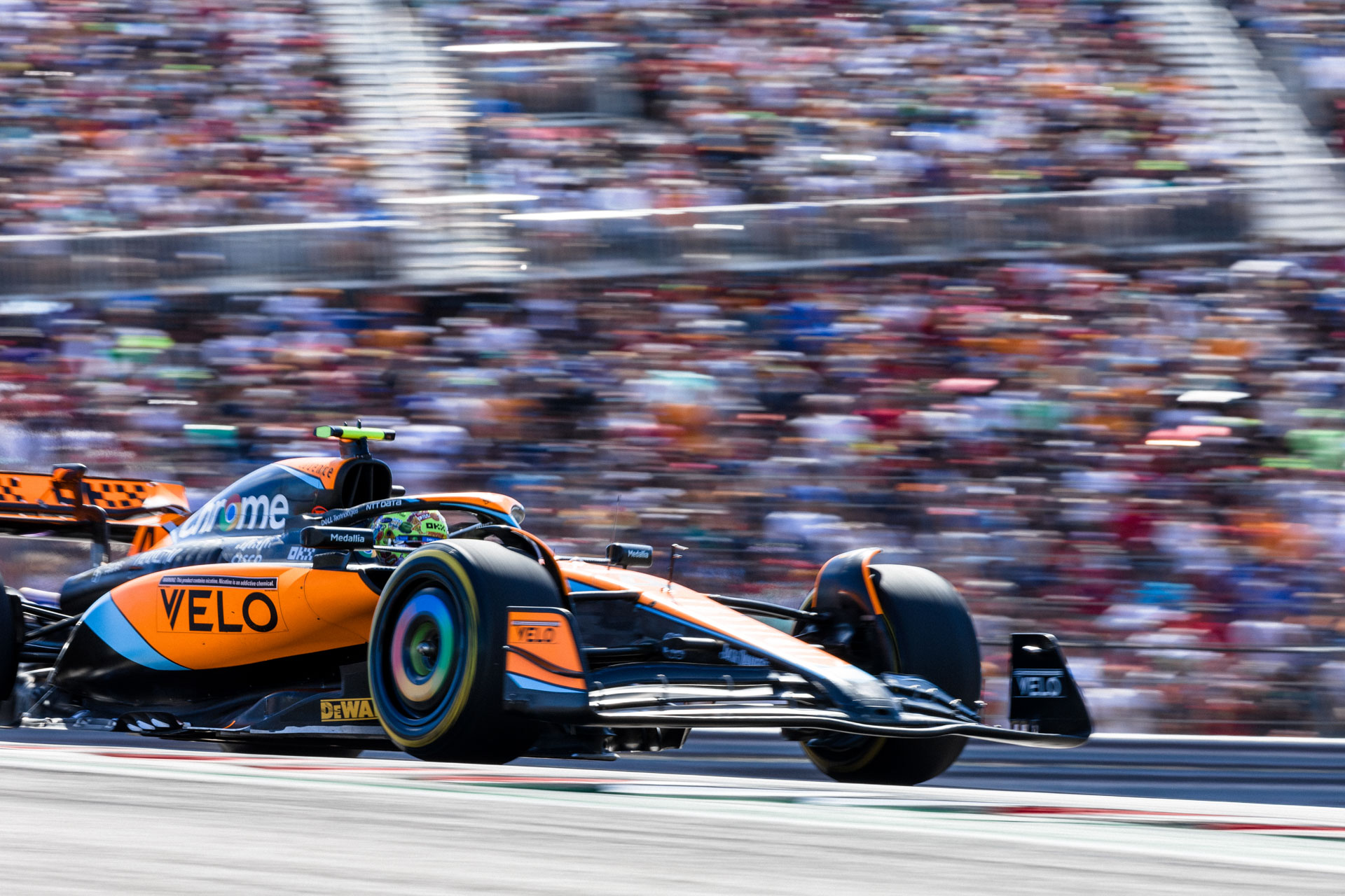 The front of an orange F1 car is just visible as it passes in front of a blurred crowd