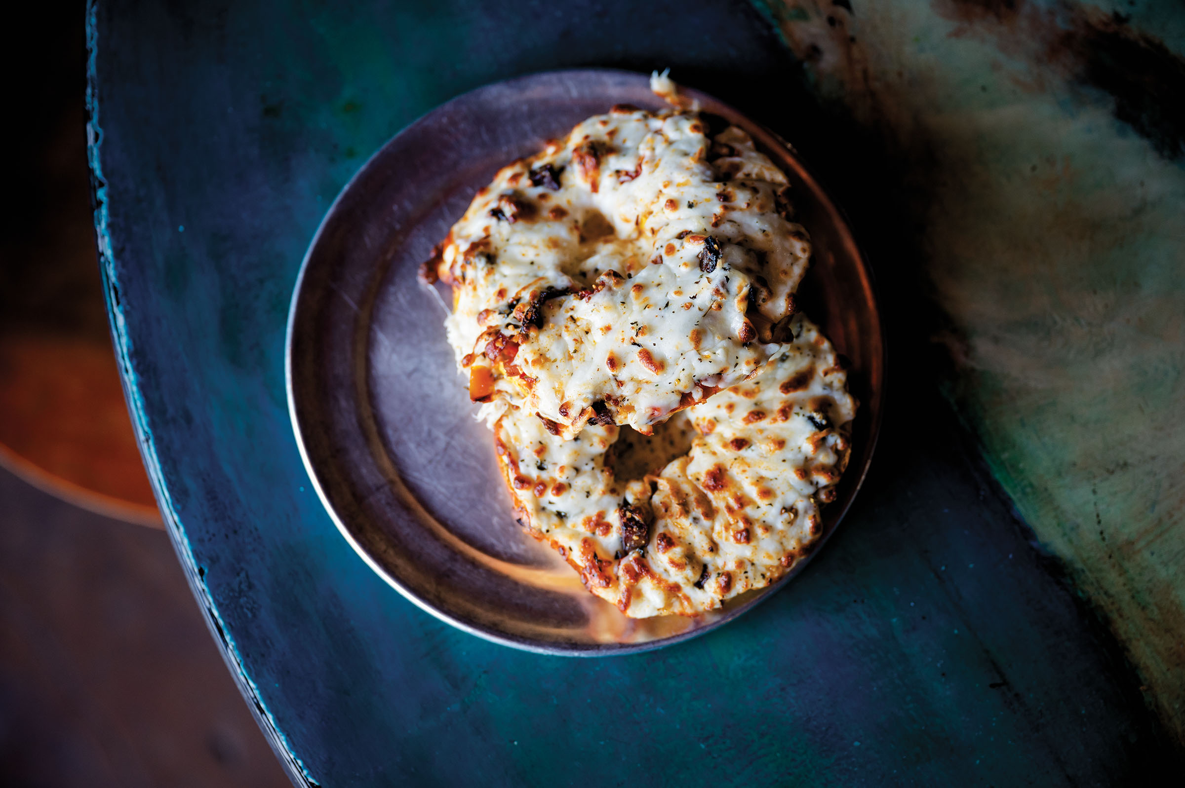 An overhead picture of a cheesy, saucy bagel on a dark blue plate