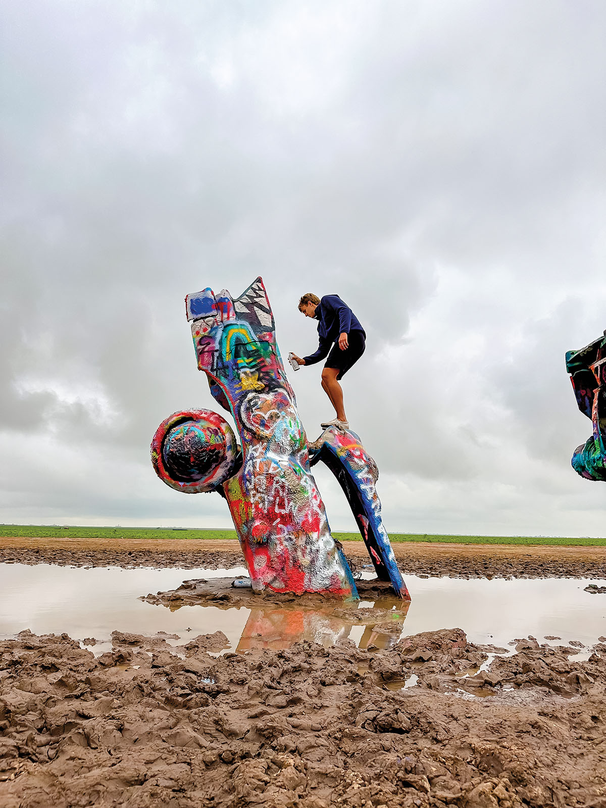 A person balances on a spraypainted car with a can of spraypaint in a muddy environment