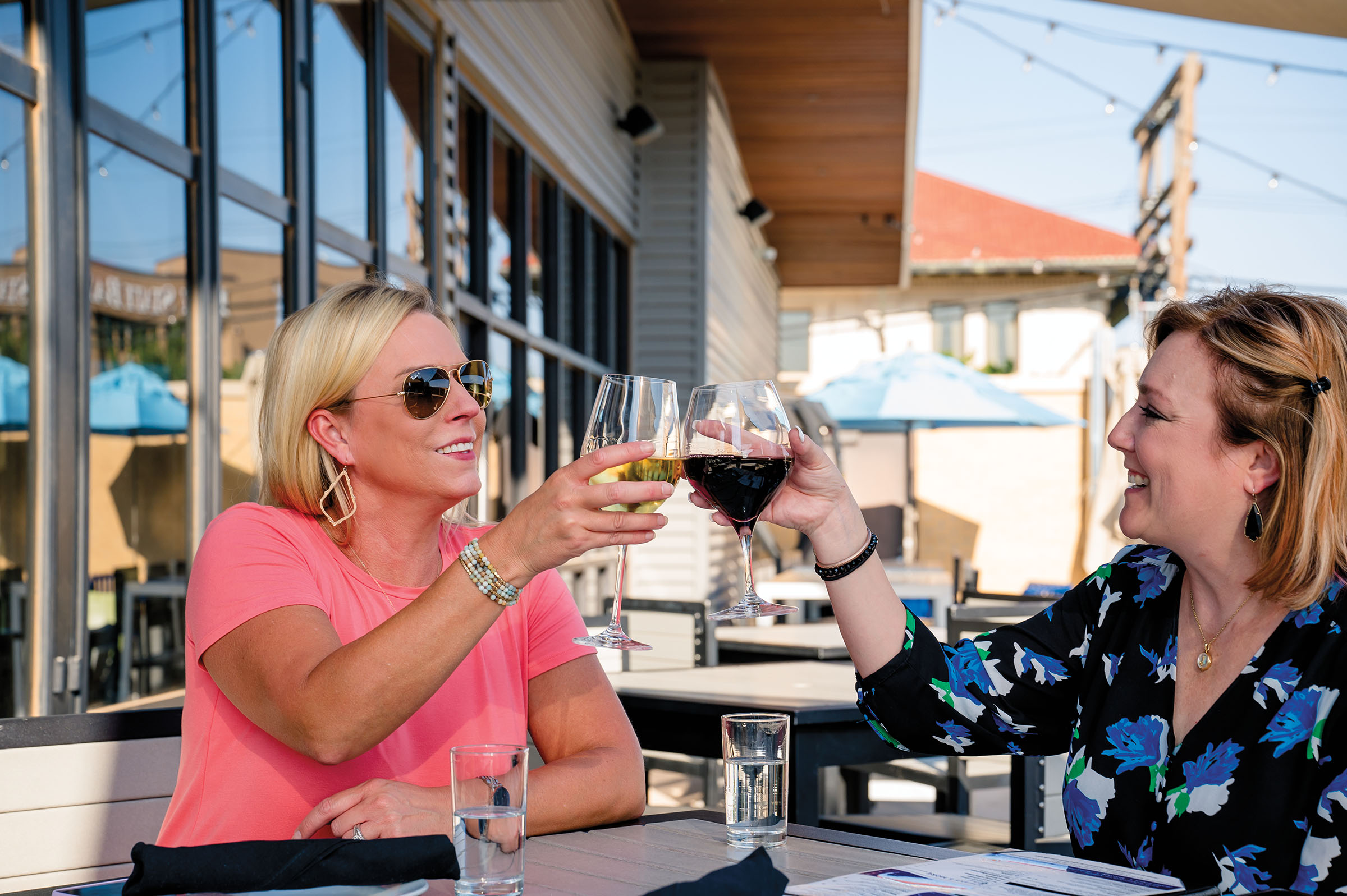 Two women cheers wine glasses while seated outside of a restaurant