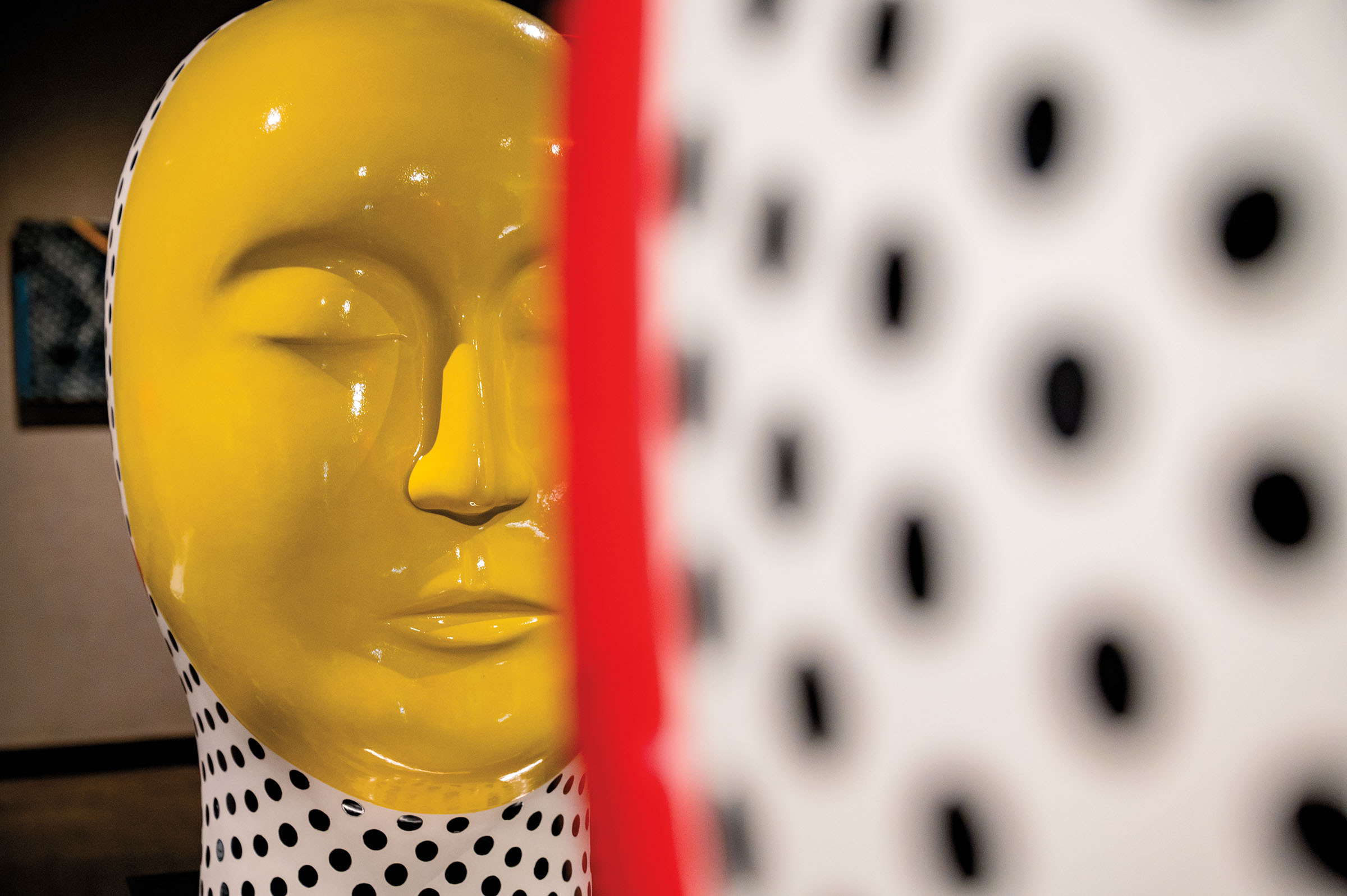 A sculpture in the shape of a yellow face sits in the background of a museum with polka dots in the foreground