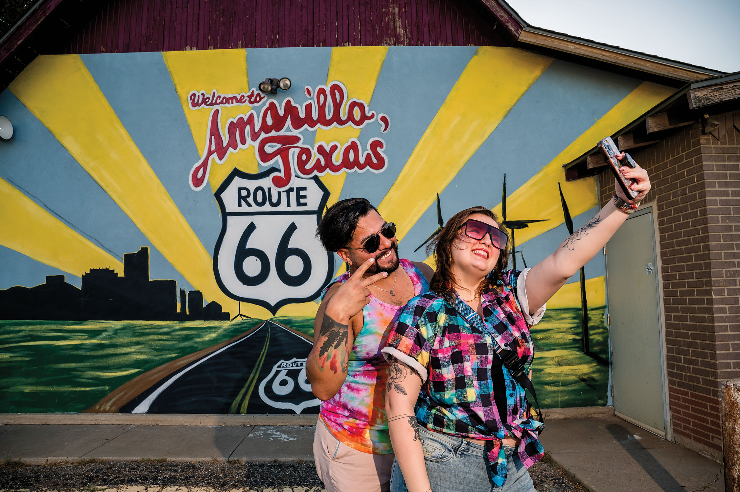 Two people in brightly-colored shirts pose for a selfie in front of a mural reading "Welcome to Amarillo, Texas"