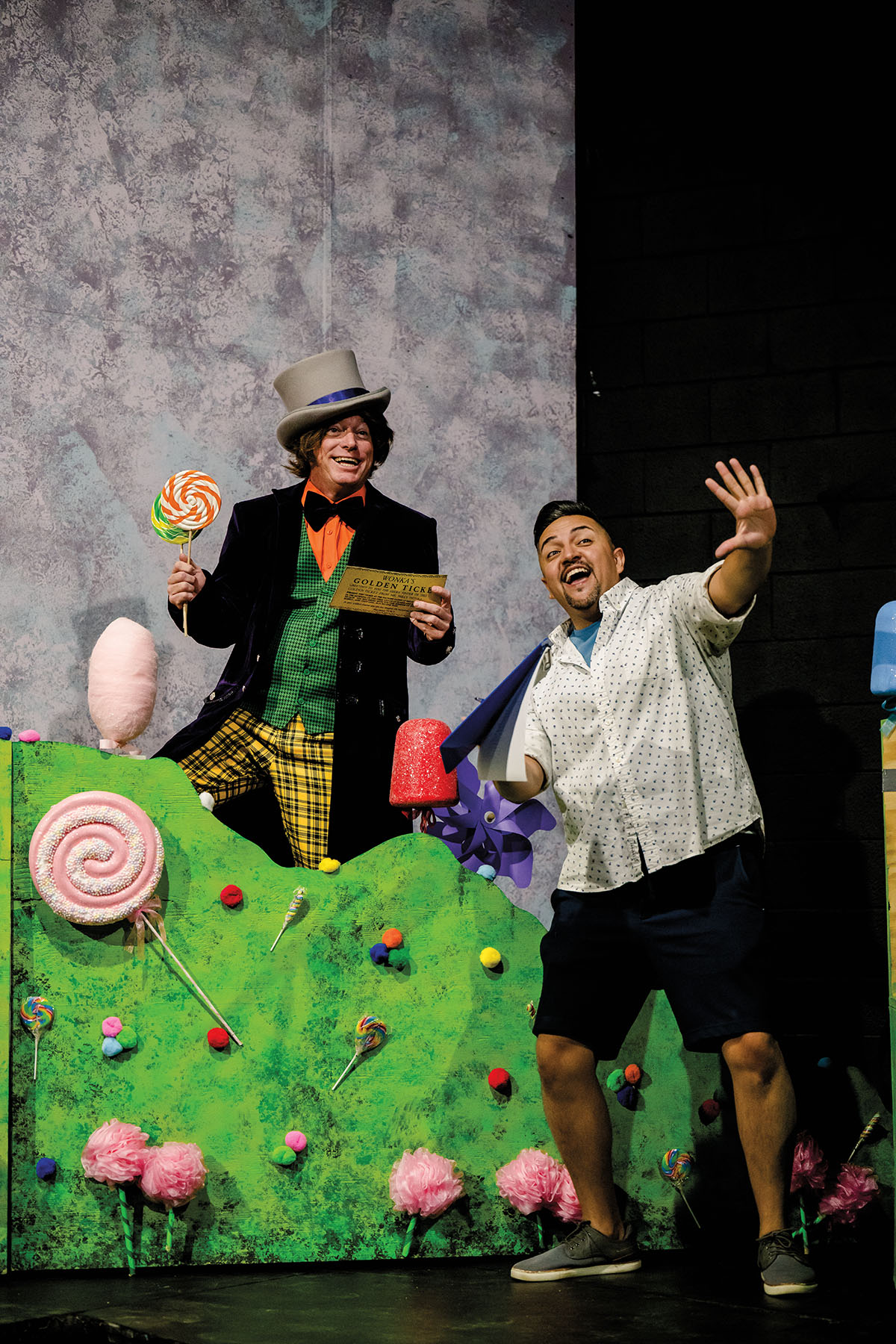 Two men, one in a brightly colored costume, rehearse under stage lights in a whimsical scene