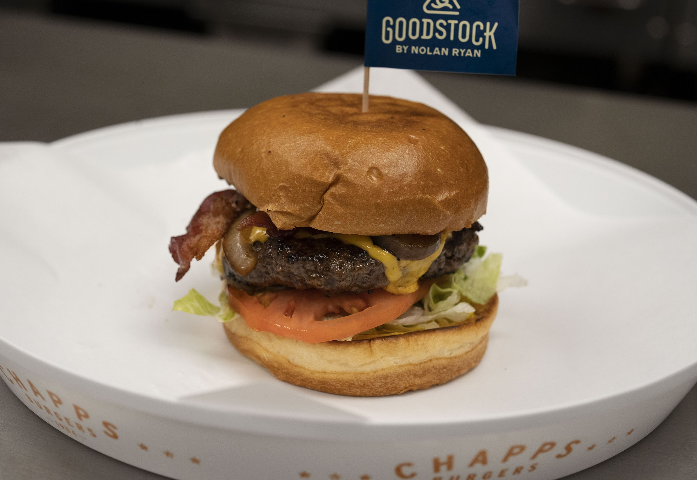 A burger with a toasted brown bun sitting on a white plate. The burger has a small blue flag sticking out of the top reading "Goodstock"
