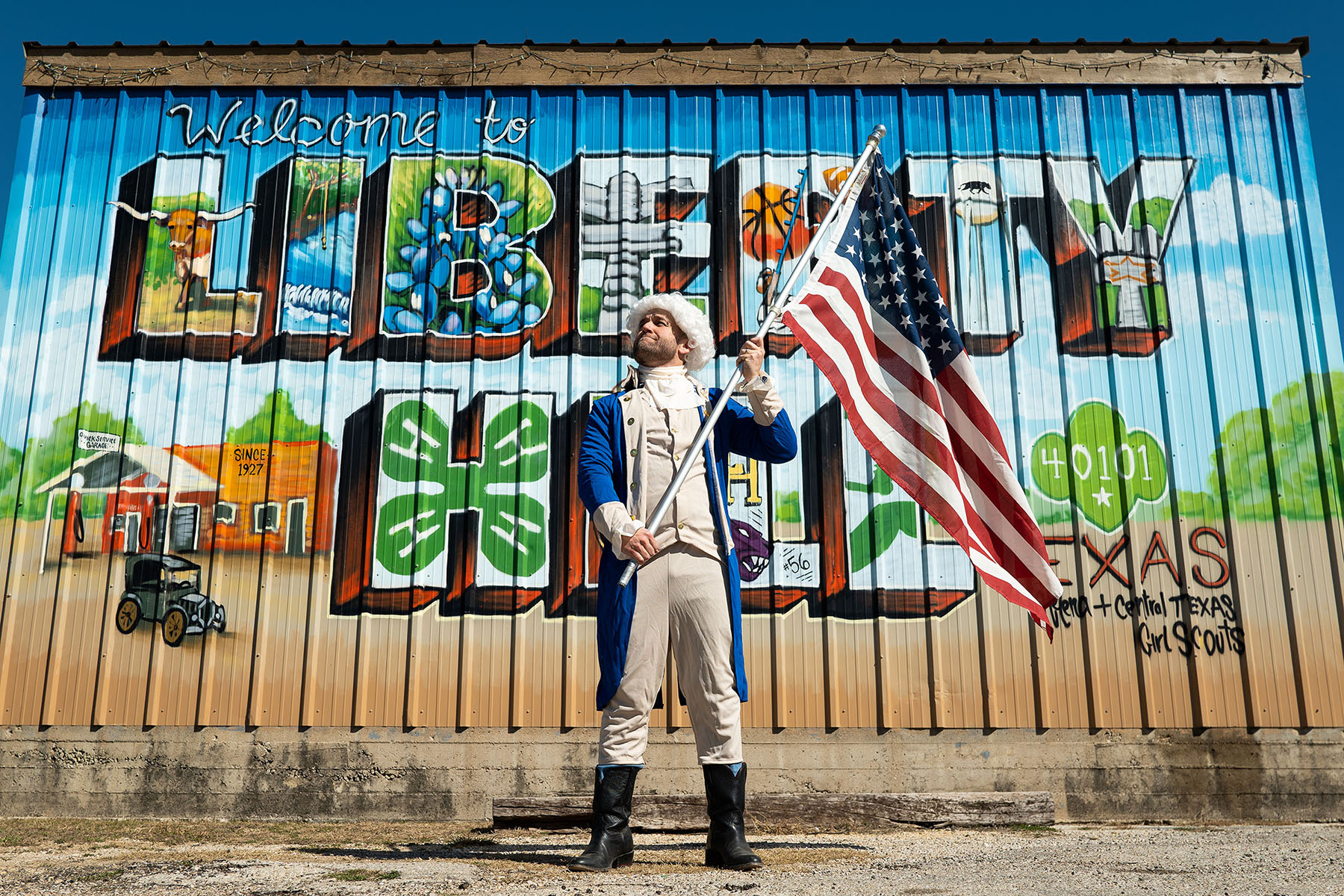 A man wearing traditional Colonial clothes stands holding an American flag in front of a mural reading "Liberty Hill"