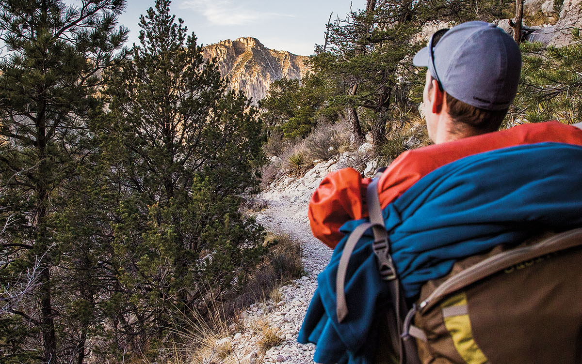 A man in a gray baseball with large hiking backpack walks along a tree-lined rock trail