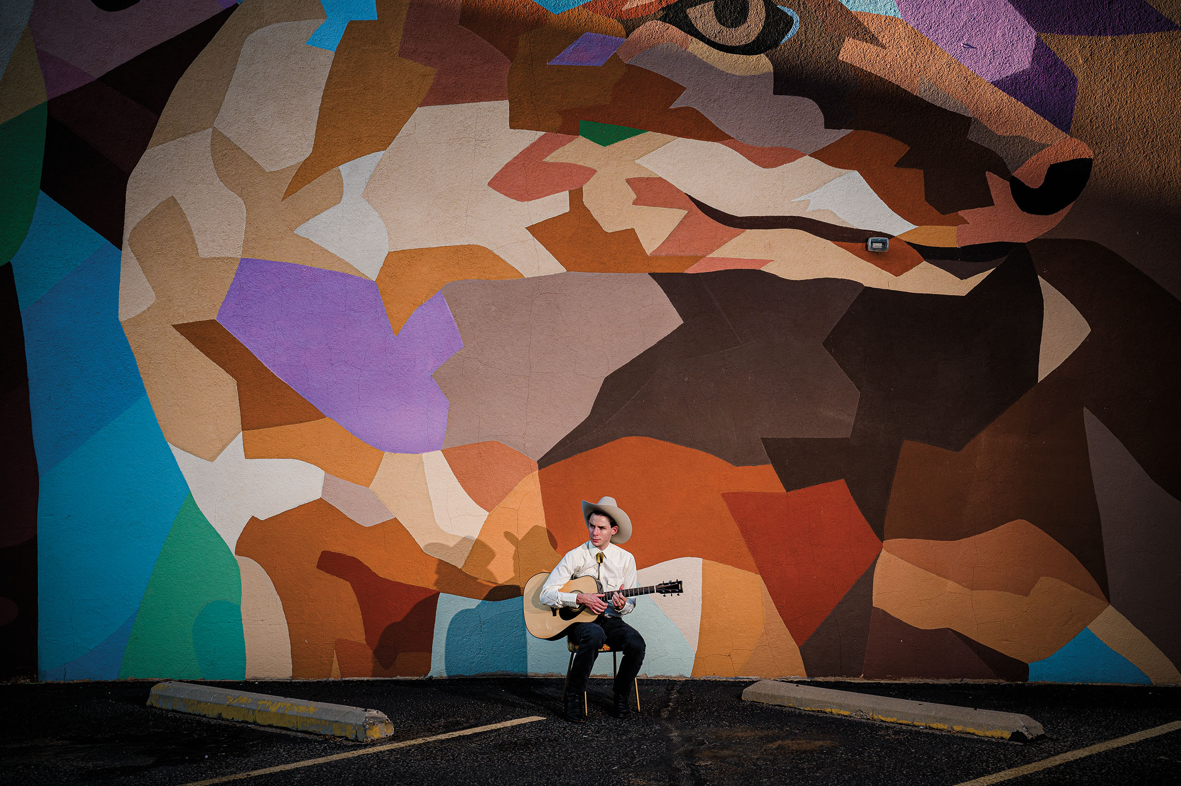 A young man wearing a cowboy hat and white shirt sits in a chair with a guitar beneath a brightly colored mural