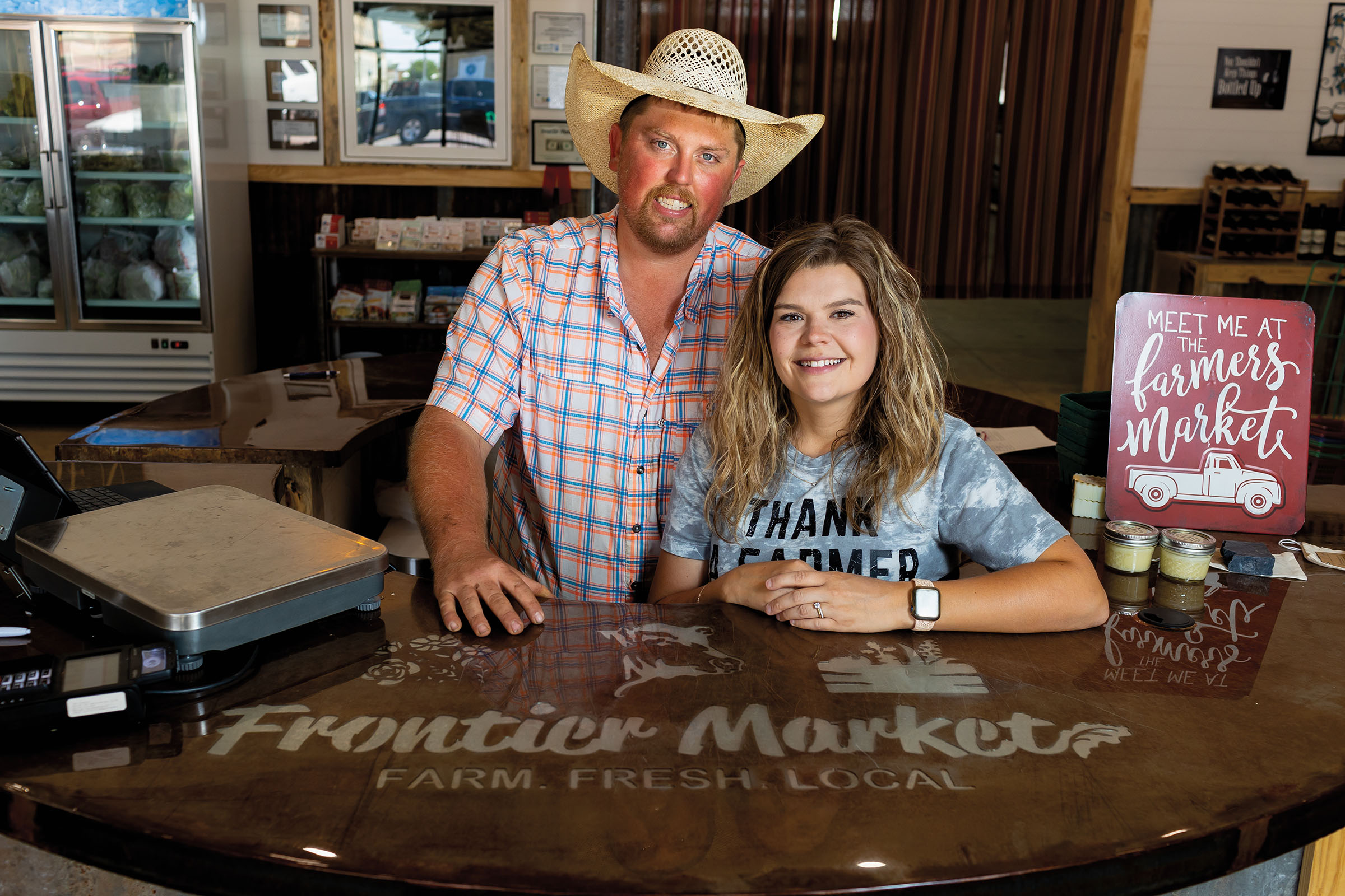 Two people, one wearing a cowboy hat, stand at a shiny countertop engraved with the words "Frontier Market. Farm. Fresh. Local."