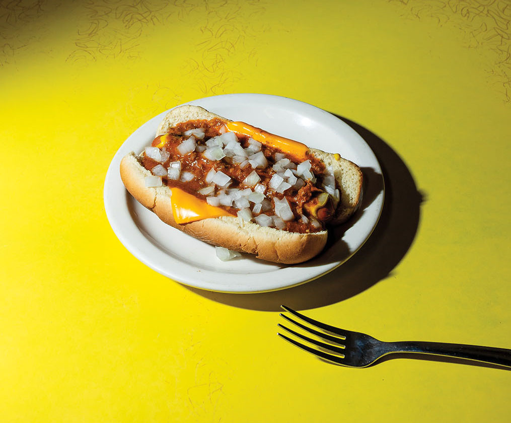 A photo of a chili dog on a white plate sitting on a yellow table