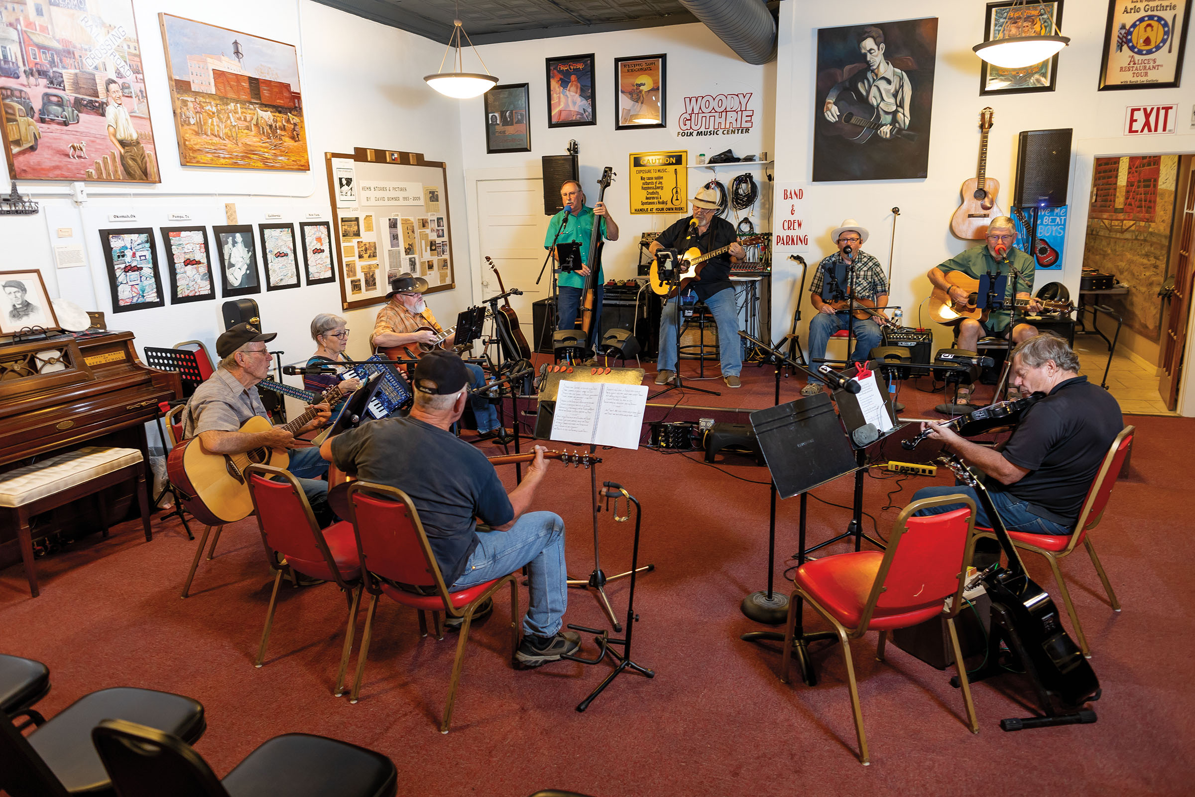 A group of people sit in red chairs on a red carpeted floor playing various instruments and singing