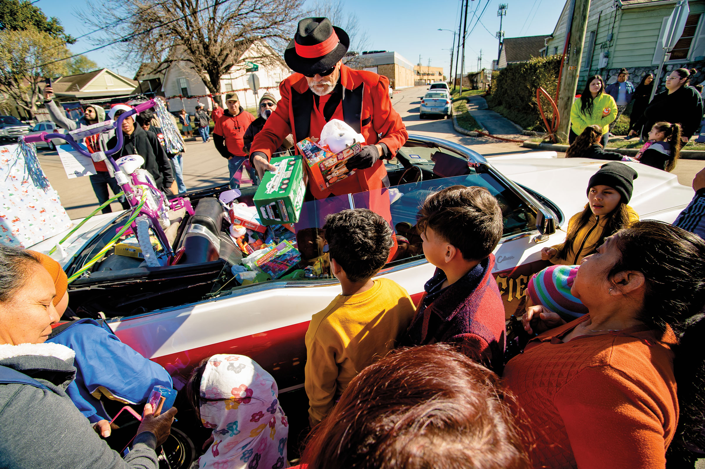 A man in a bright red zoot suit and black hat hands out presents to children from the back of a red convertable car