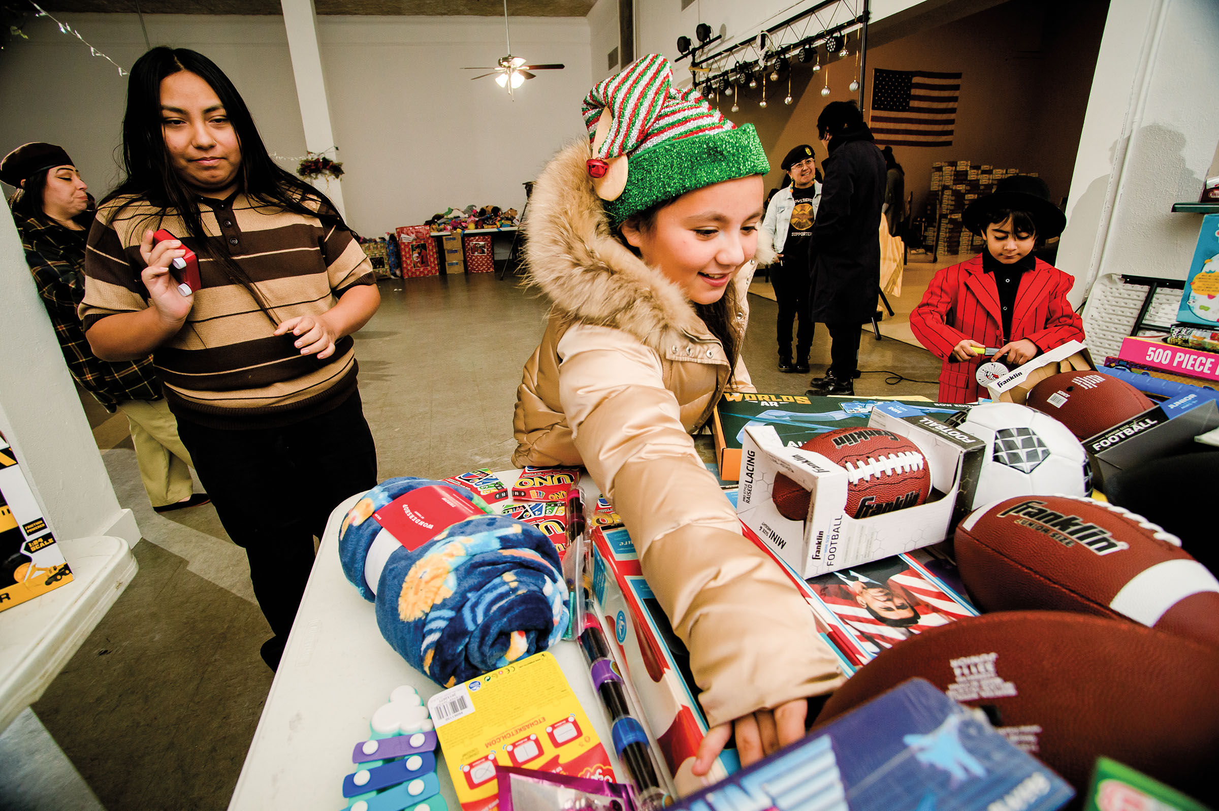 A young person wearing a striped elf hat reaches over a table full of toys and other gifts