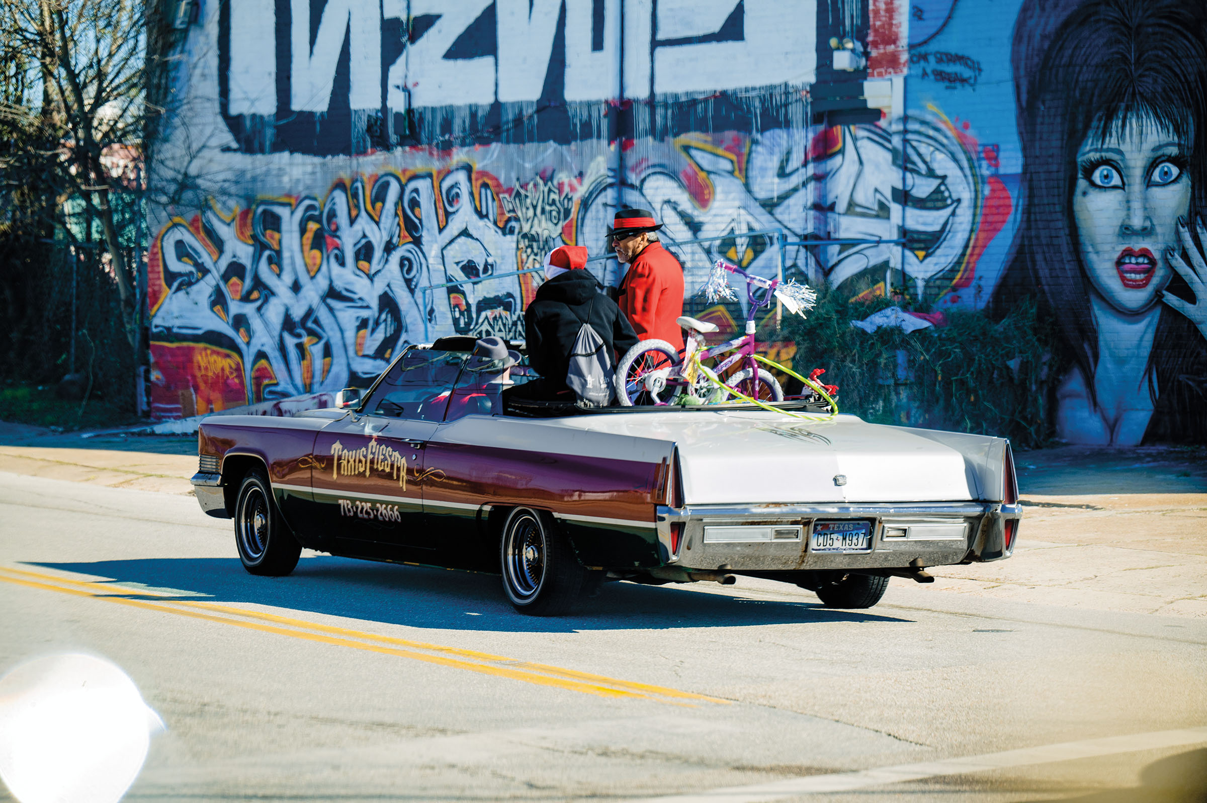 Pancho Claus, in his typical red zoot suit, sits in an open convertable as it drives in front of a brightly colored mural with spray painted graffiti