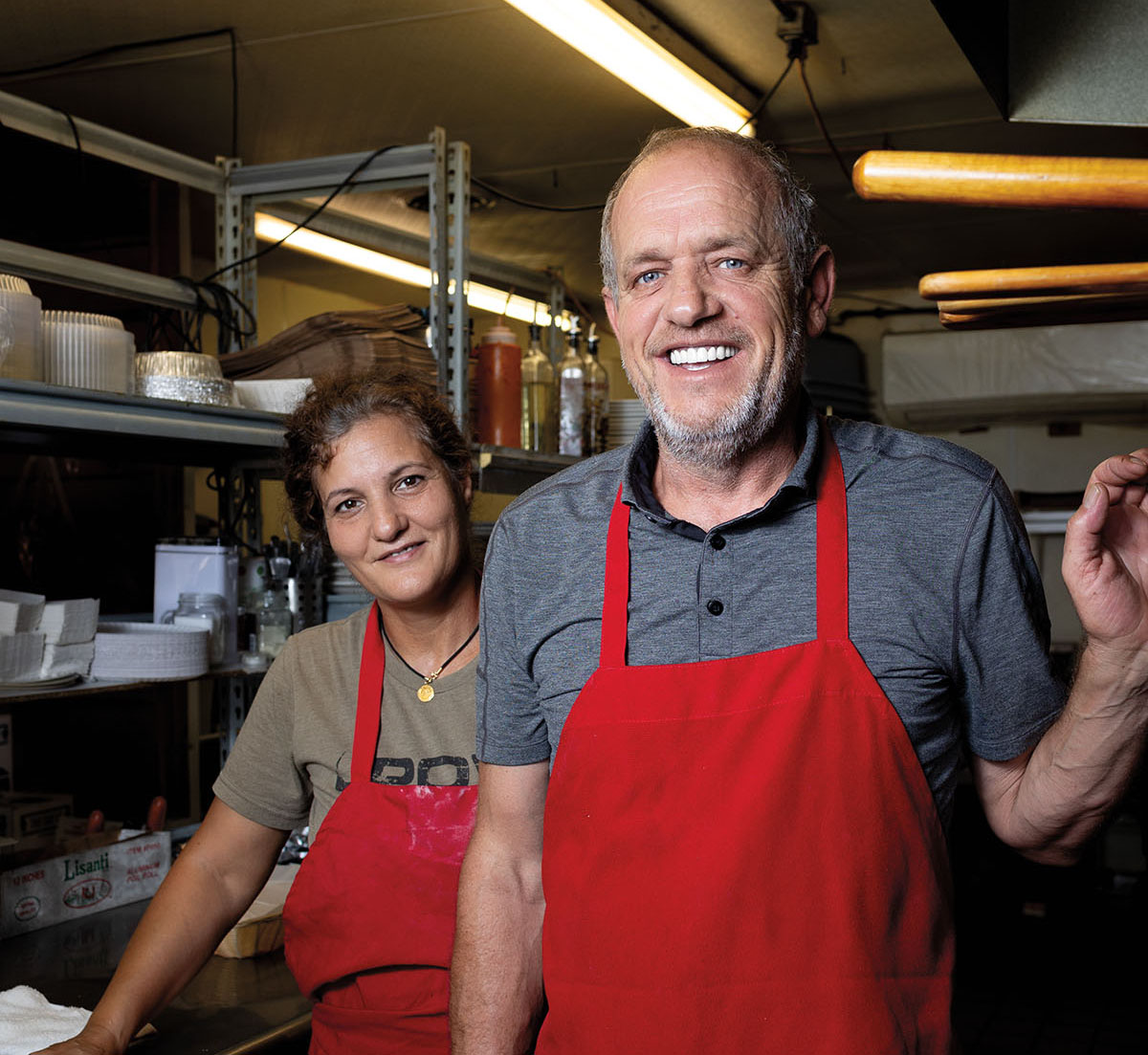 Two people in red aprons smile in a kitchen setting with fluorescent lights