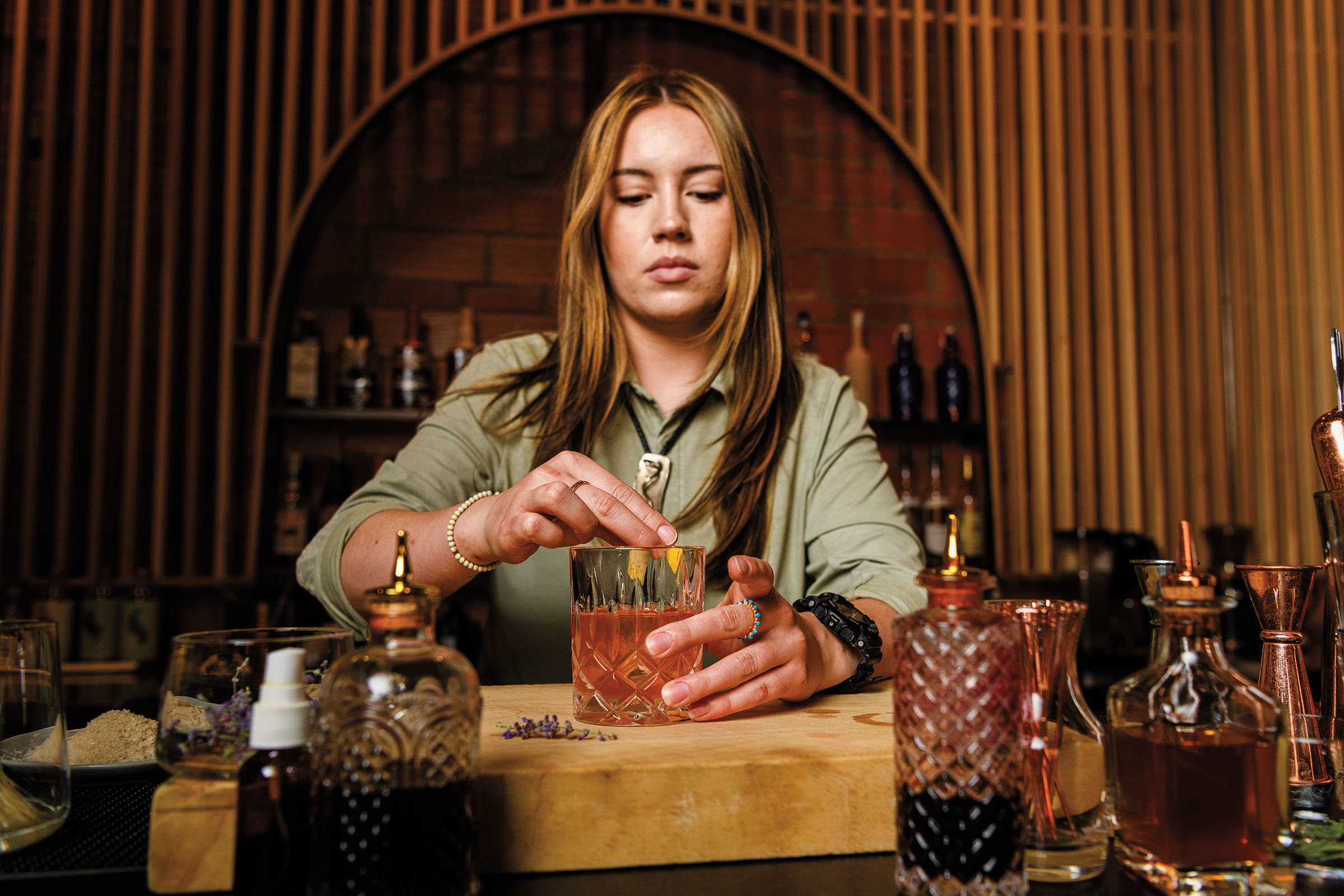 A woman wearing a green shirt makes a craft cocktail in a Collins glass in a rich wood bar