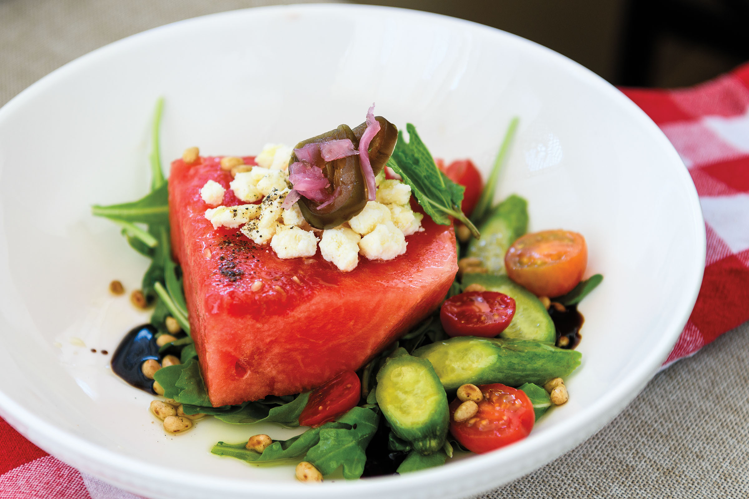 A white bowl filled with numerous vegetables and a large slice of watermelon topped with cheese and olives