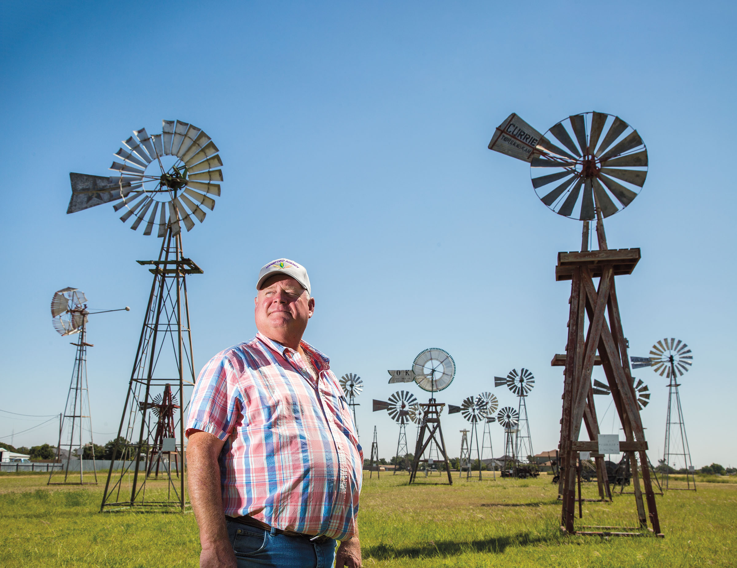 A man in a striped shirt and baseball cap stands in a field with numerous wooden old-fashioned windmills