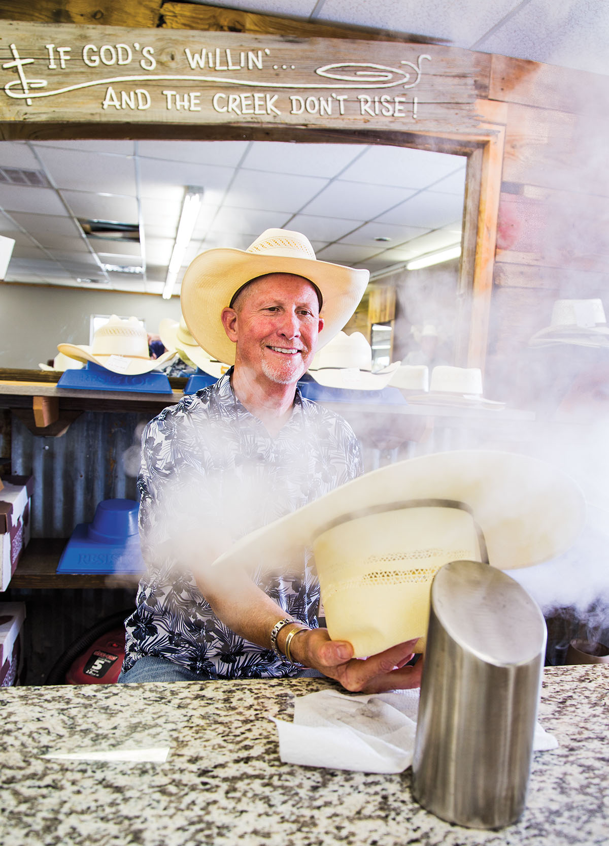 A man in a cowboy hat steams a tan hat on a silver table