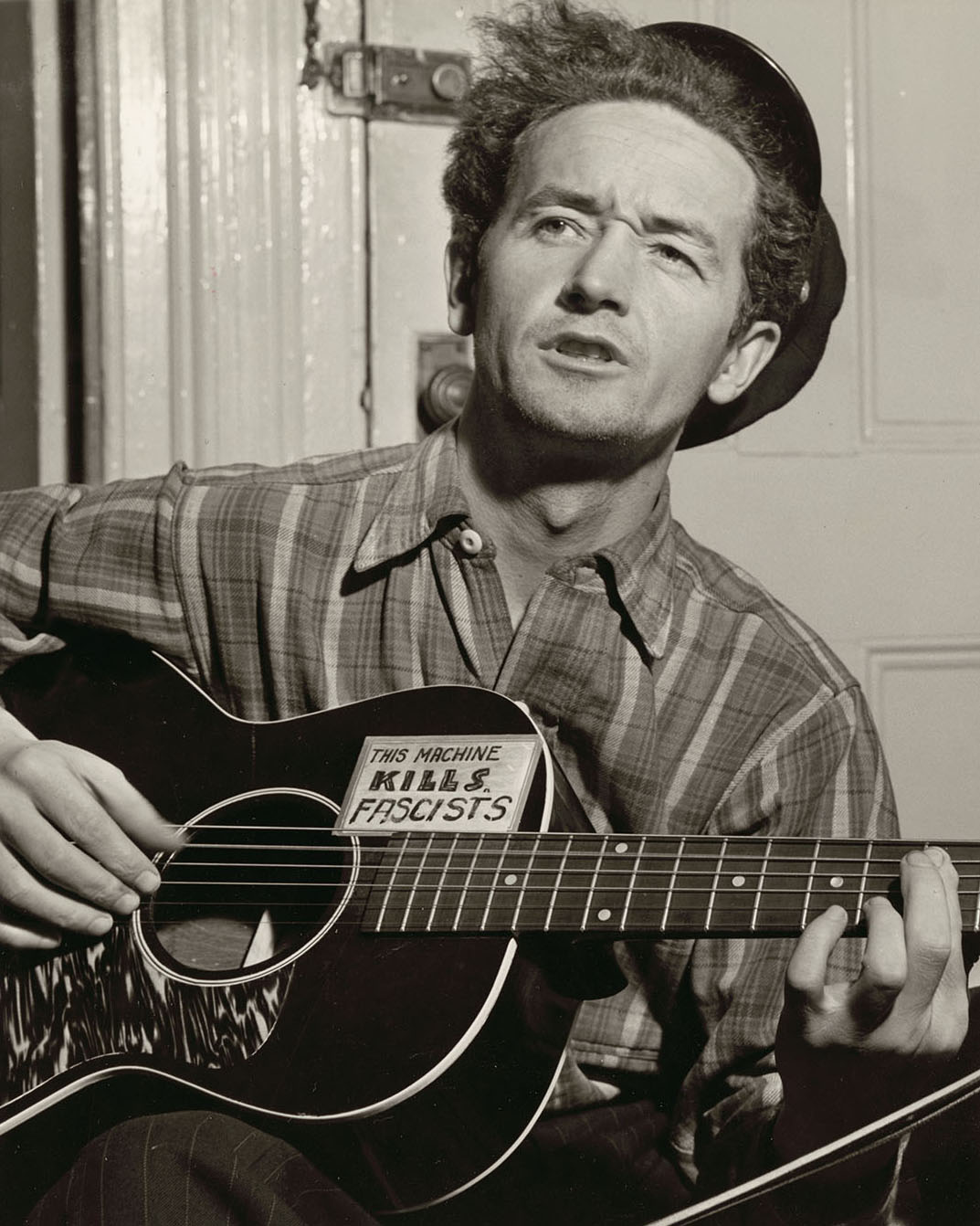 A sepia-tone photograph of a man wearing a striped shirt and small hat. He holds a guitar with a small note on it reading "This machine kills fascists"