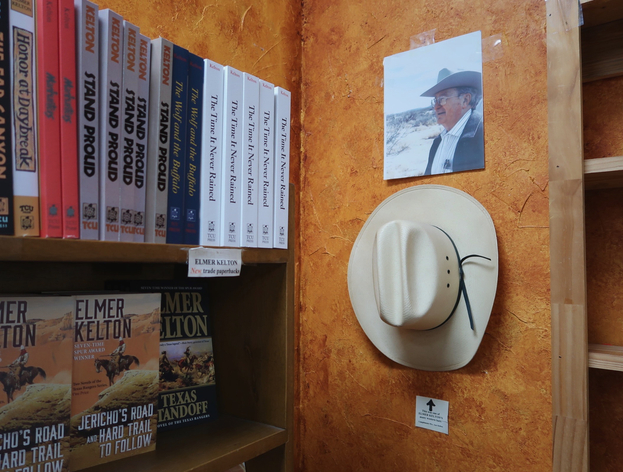 Books sit on shelves against a wall bext to a hanging cowboy hat and photo of author Elmer Kelton