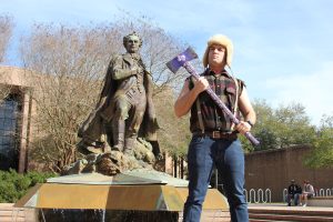 A man wearing a cutoff shirt and wool hat stands holding a large purple axe in front of a statue of Stephen F. Austin