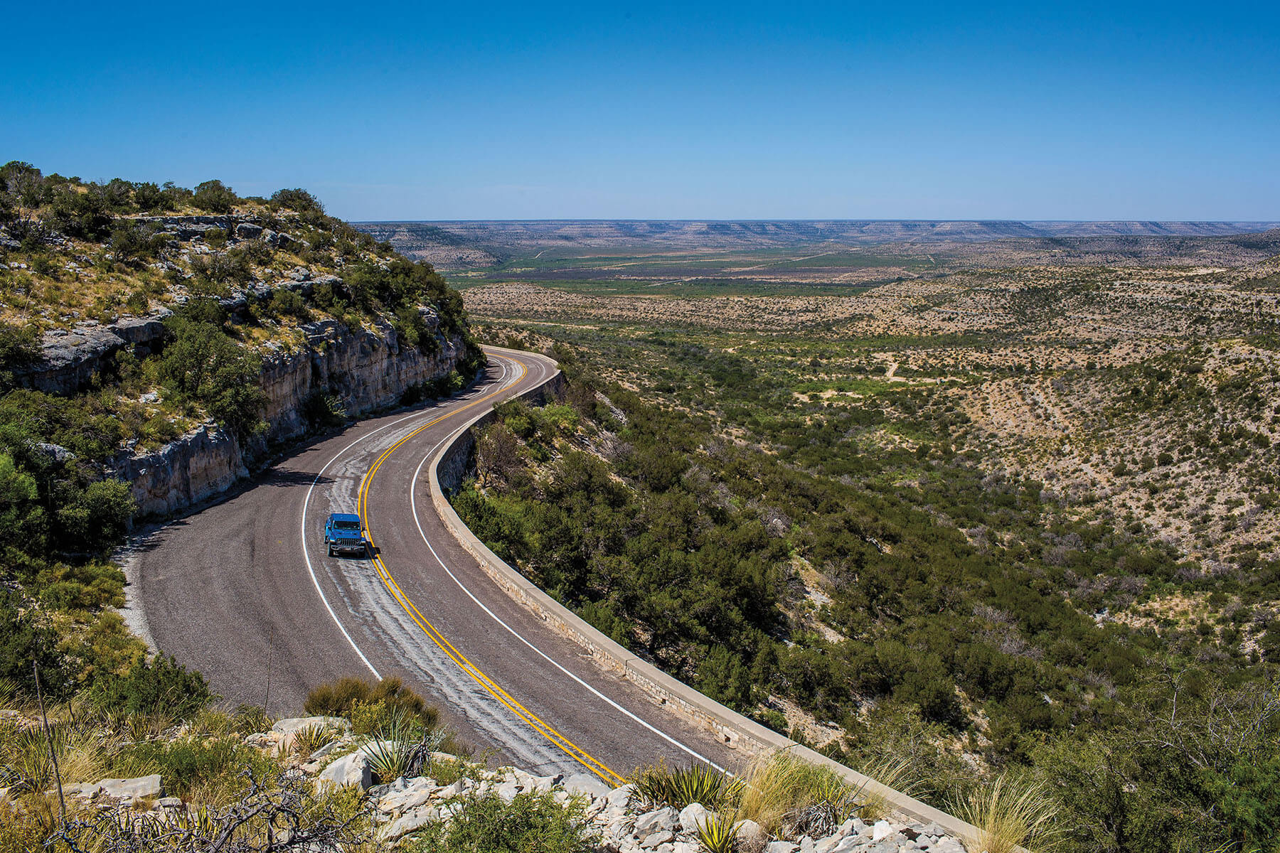 A lone blue car traverses a two-lane paved highway through desert land with short scrub