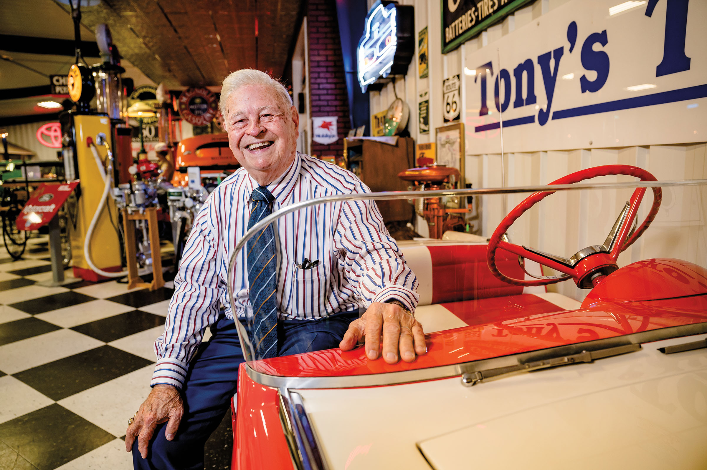 An old man in a striped shirt and tie sits in a red classic car laughing