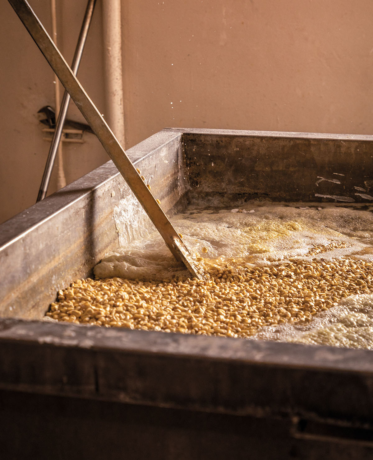 A wooden instrument is used to stir a large vat of boiling corn