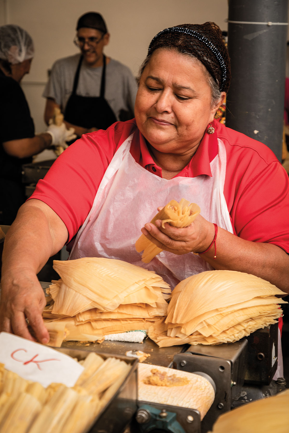 A woman wearing an apron and red shirt holds corn husks used in making tamales
