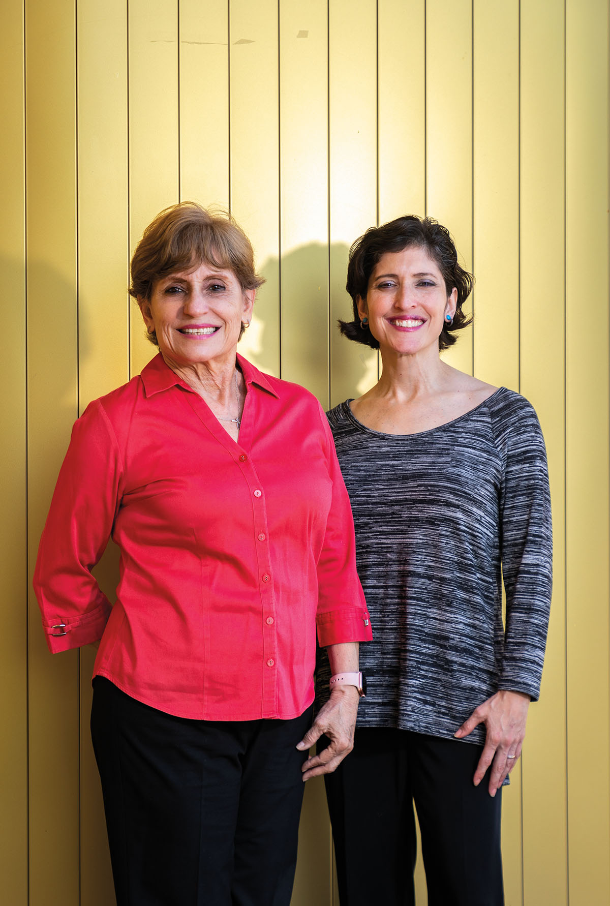 Two women stand in front of a yellow background 