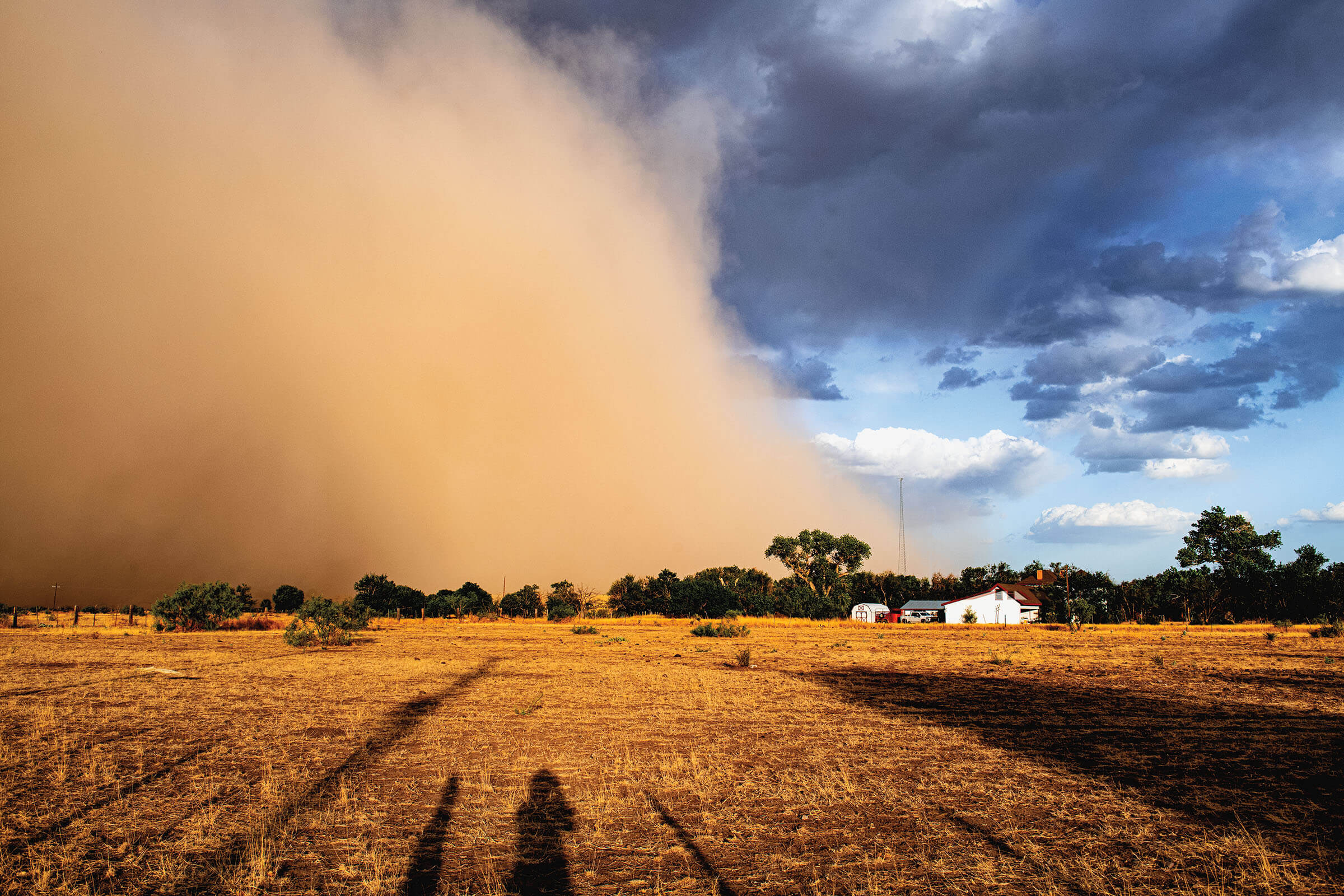 A golden dust storm moves into the frame from left, where a small farmhouse and scrub grass is visible beneath a cloudy blue sky