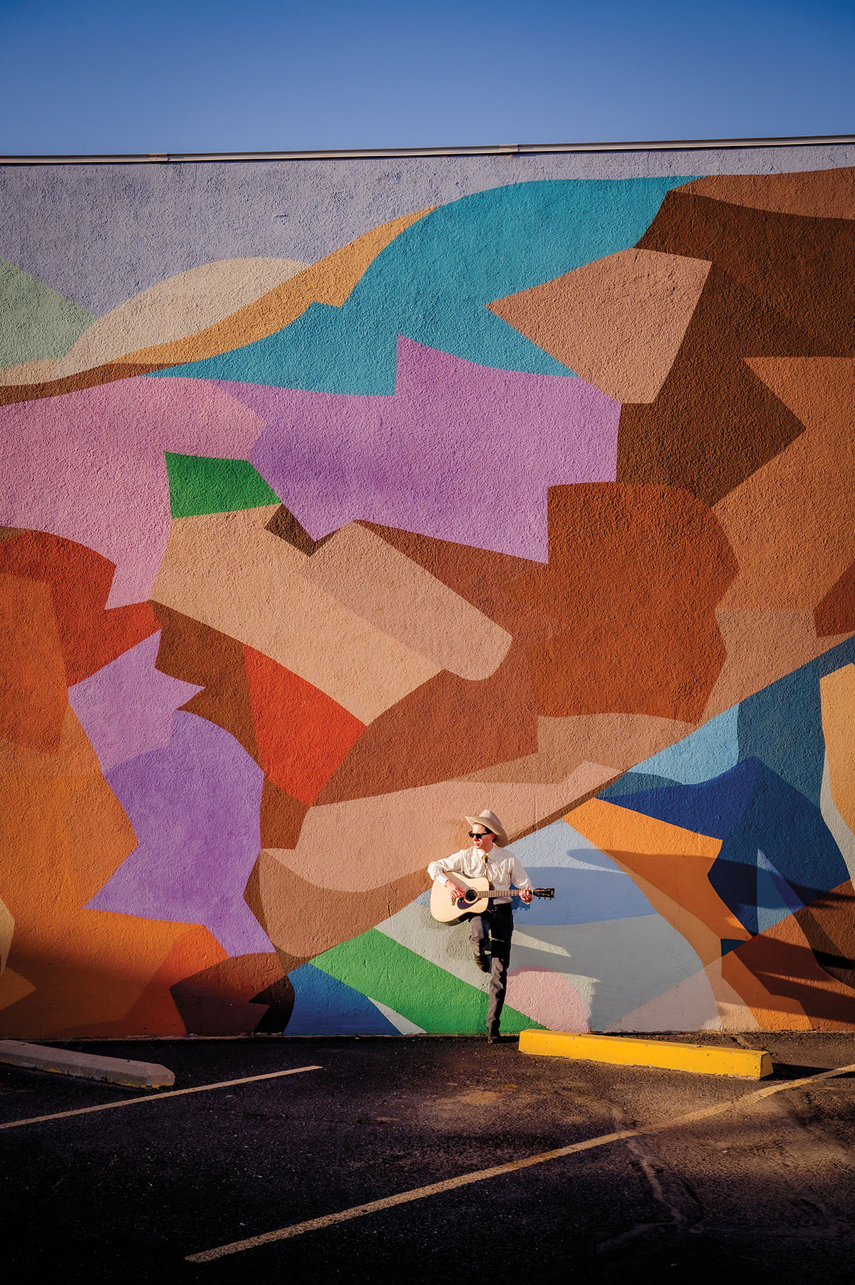 A man in a white button-down shirt and cowboy hat leans against a brightly-colored mural