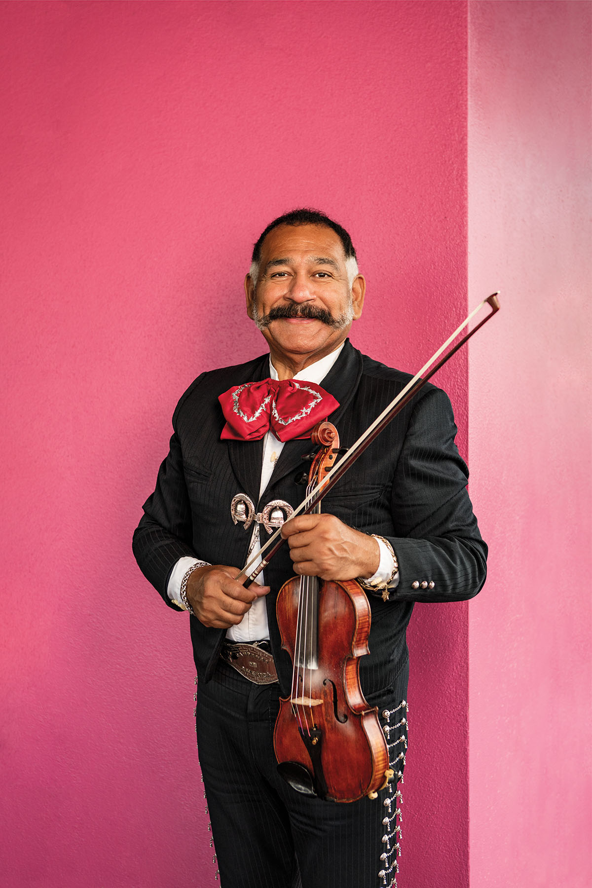 A man in a traditional Mariachi uniform stands in front of a bright pink wall
