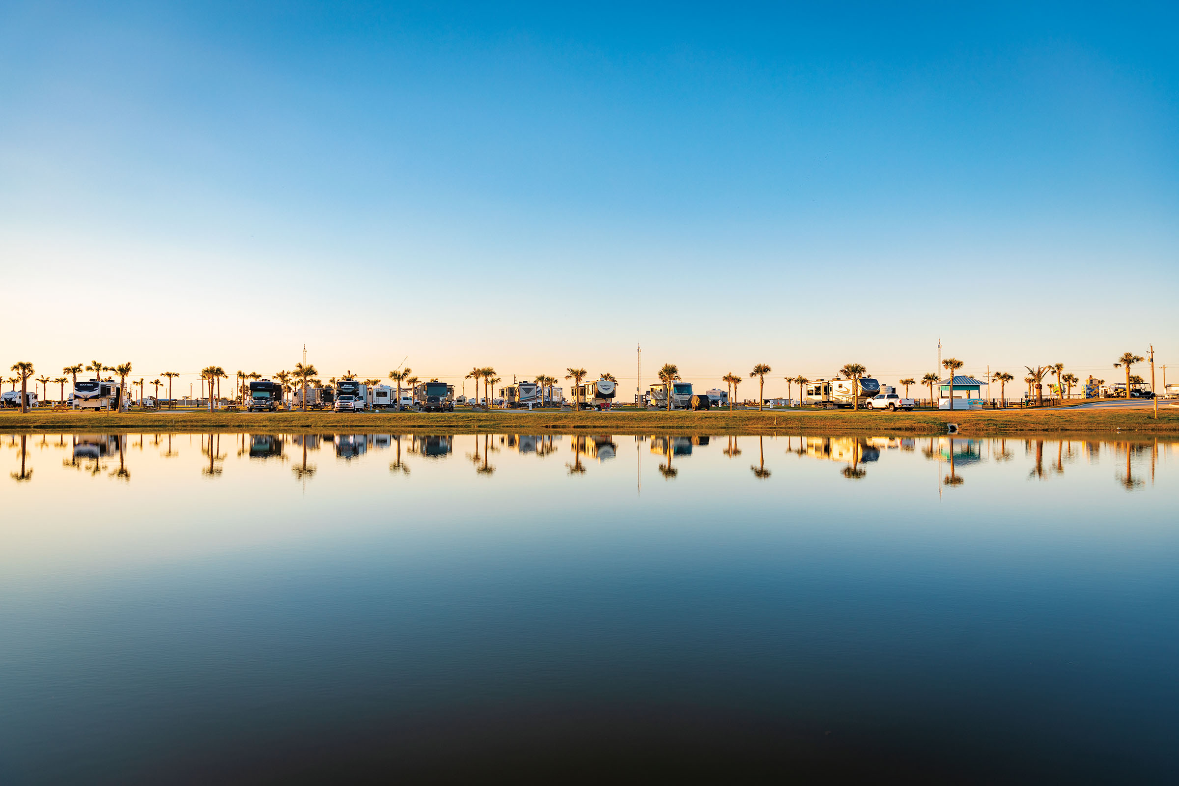 A group of RVs line up along still flat clear water in an RV park