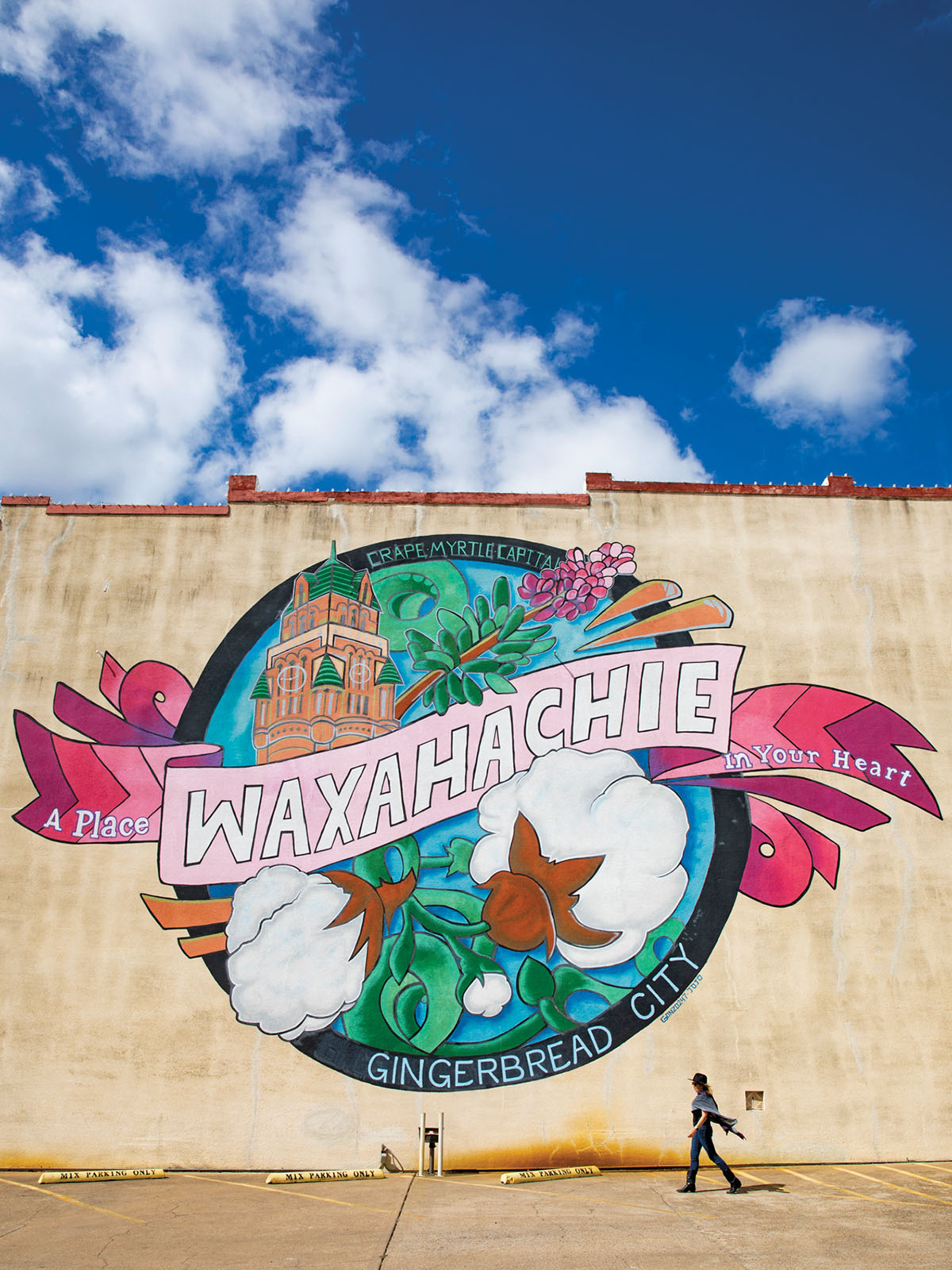 A person walks in front of a grey concrete building with a brightly-colored mural reading 'Waxahachie Gingerbread City' on the front