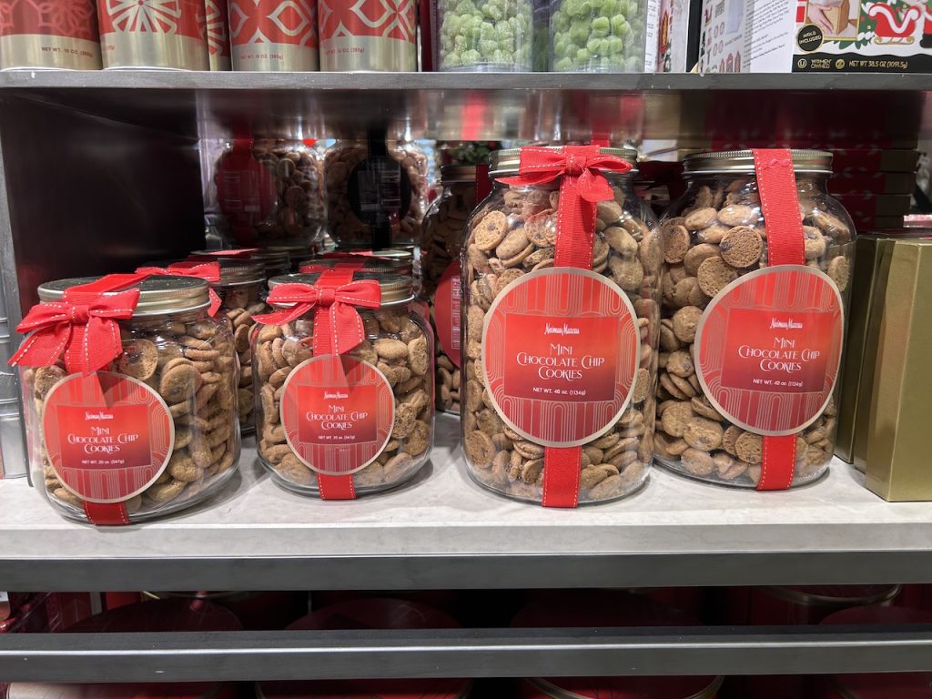 Mini chocolate chip cookies in small and large glass jars decorated red ribbons are on display on a shelf. 