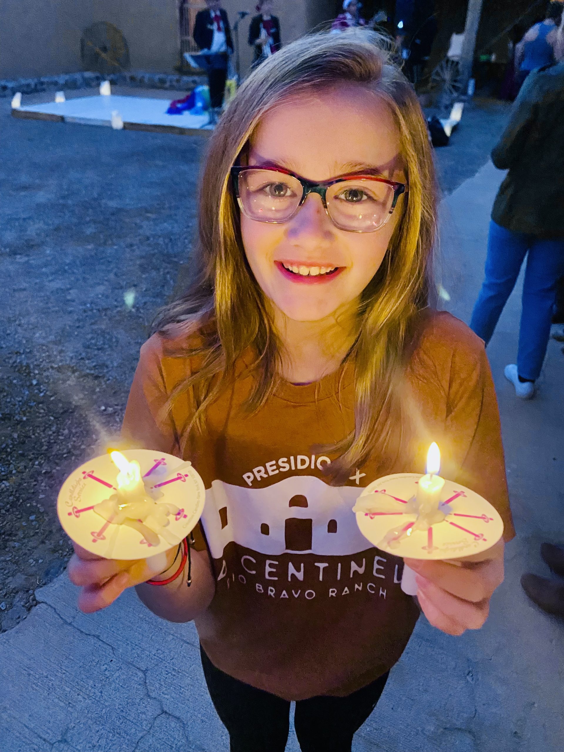A young girl wearing glasses and a Presidio Texas T-shirt holds two luminarias and smiles for the camera. 