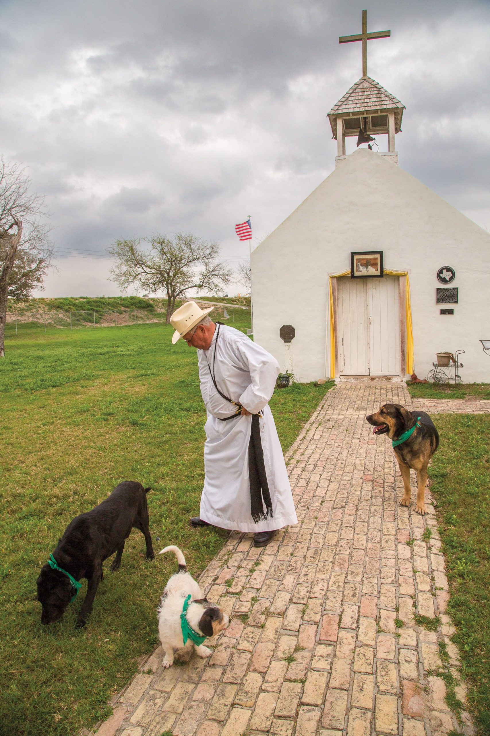 A man in a white robe and cowboy hat stands by a small white chapel with a cross on top.
