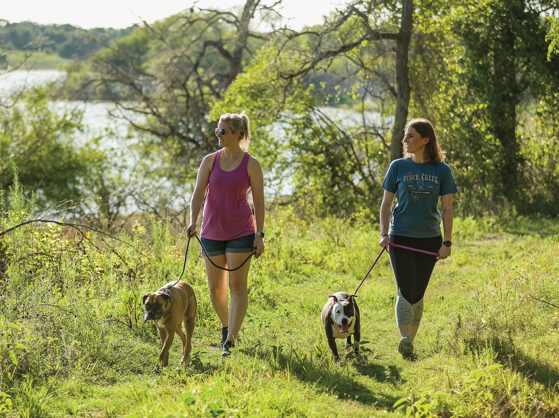Two women walk dogs down a green trail.