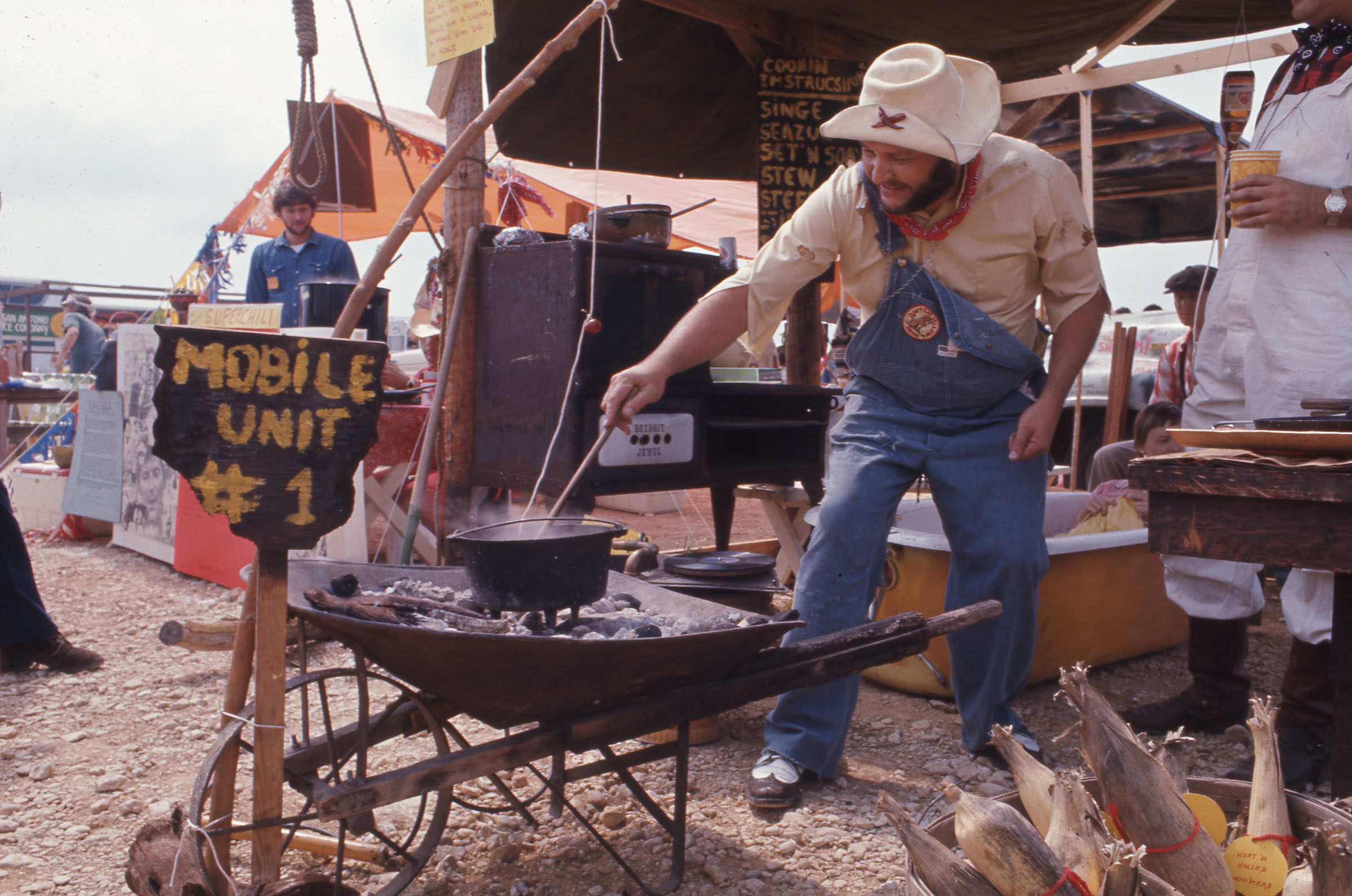A vintage photo of a man in a cowboy hat working over a fire and pot of chili