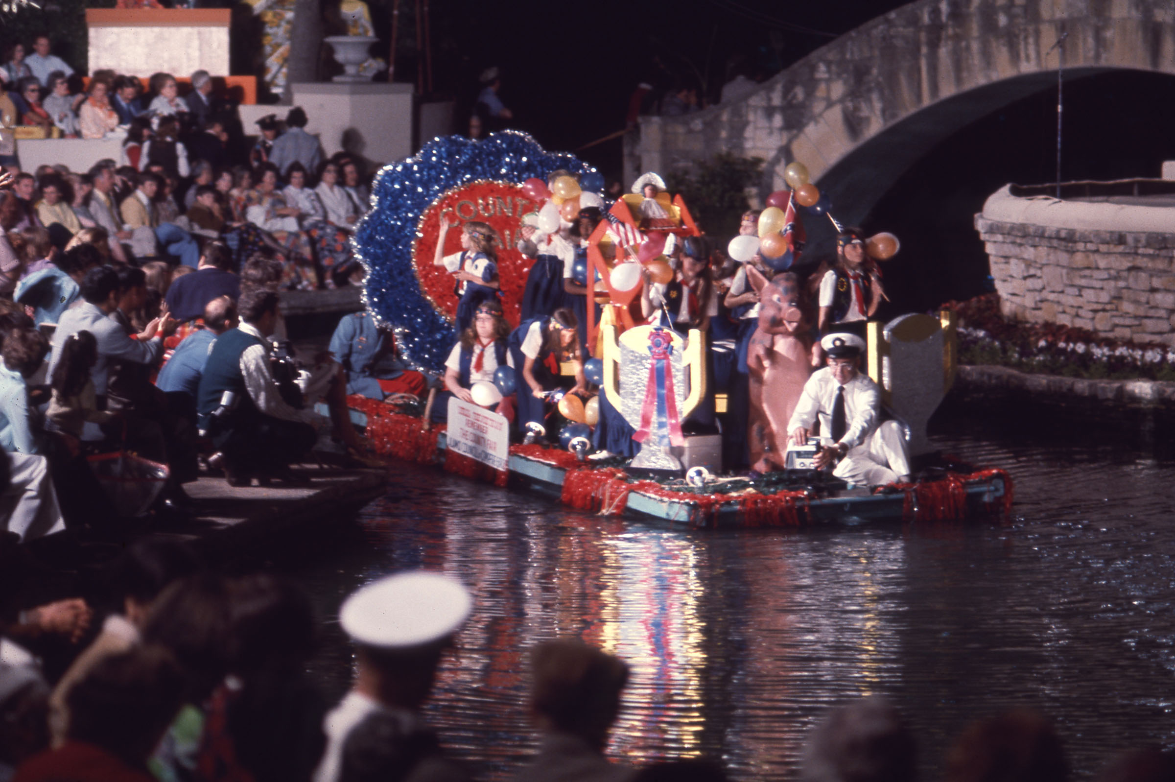A float on water with people in large costumes in front of a crowd of onlookers