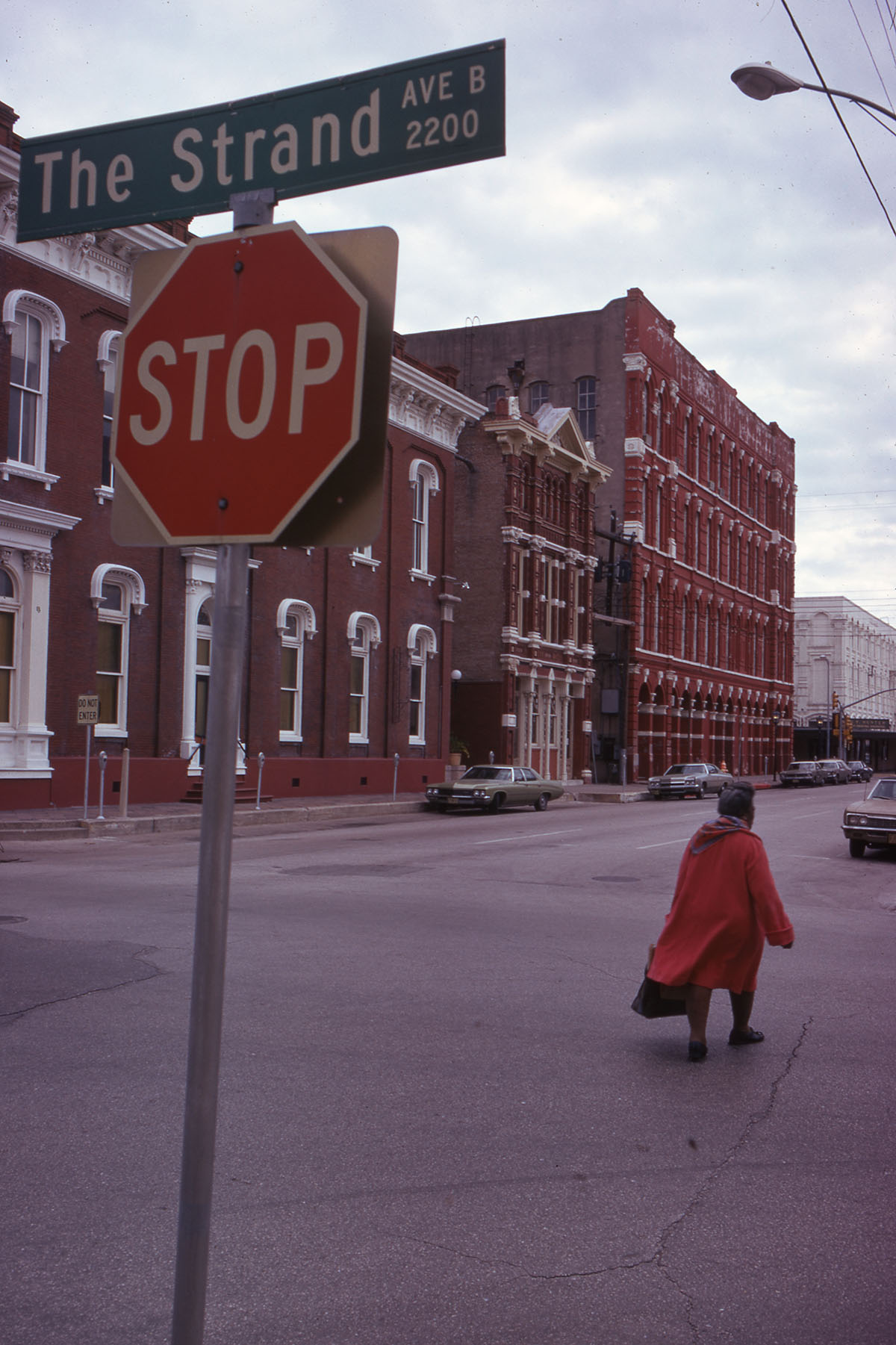 A person in a long red coat walks along a roadway under a street sign reading 