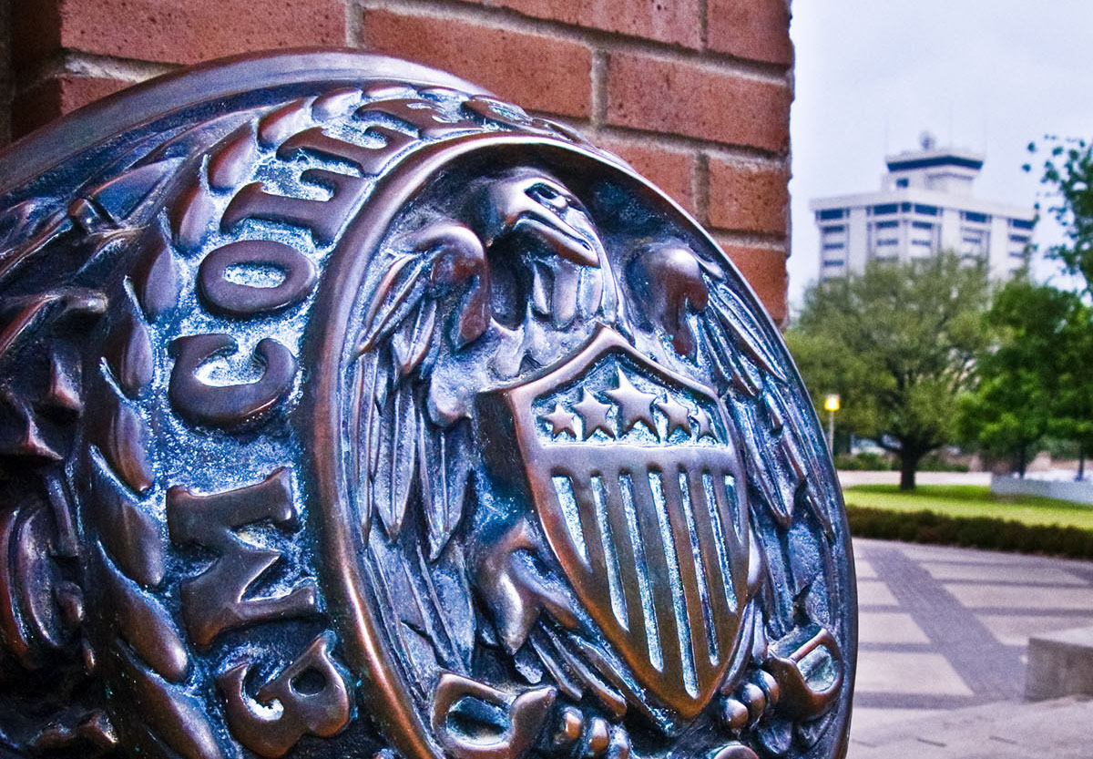 A cast bronze emblem of an eagle holding a shield. A brick wall is behind.