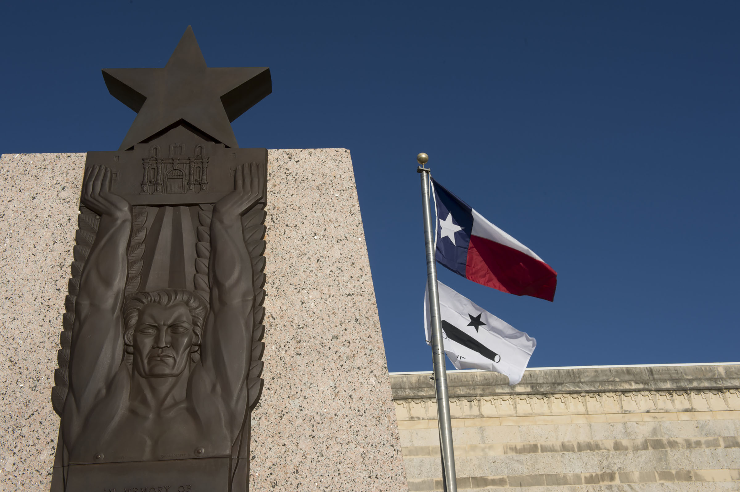 A Texas flag flies above a light-colored stone wall.